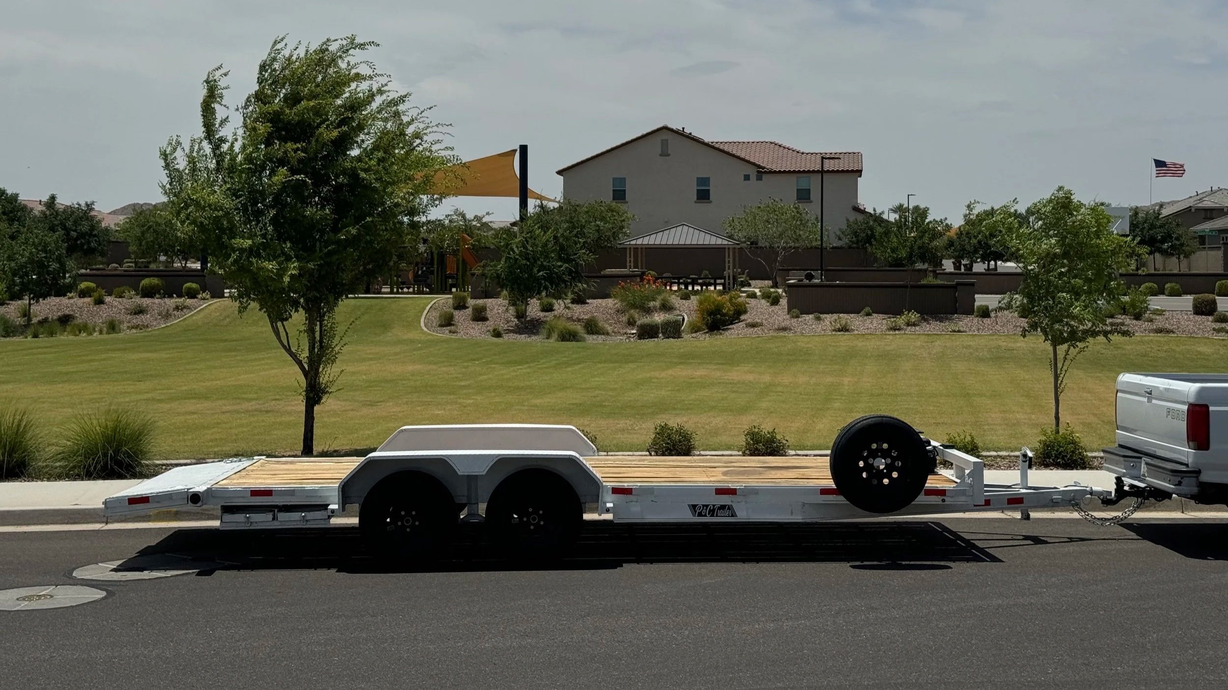 A flatbed trailer parked on the street with a tow hitch connected, water tank, and spare tire, with a suburban neighborhood, trees, a house, and an American flag in the background.