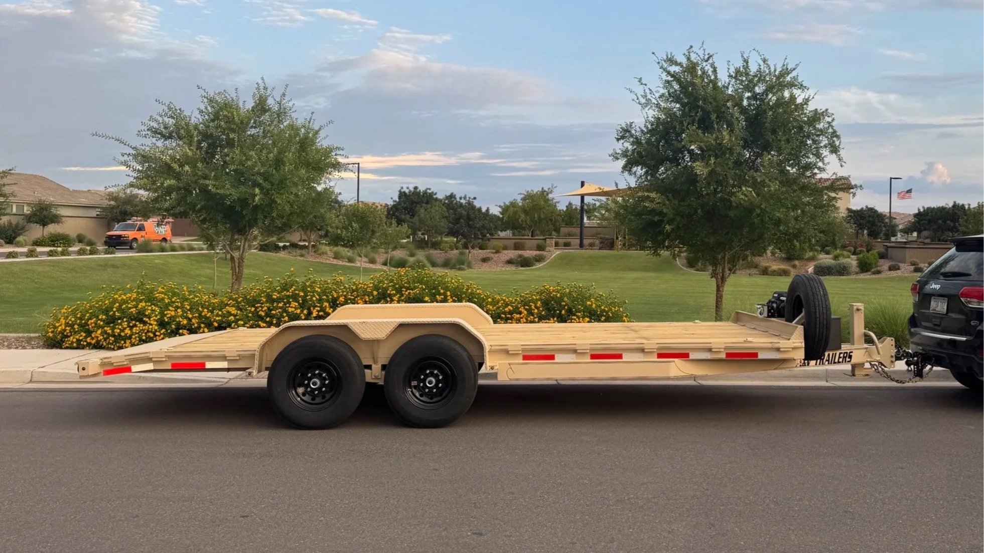 A beige flatbed trailer attached to a black Jeep SUV parked on the street with a large flower bed and green trees in the background.