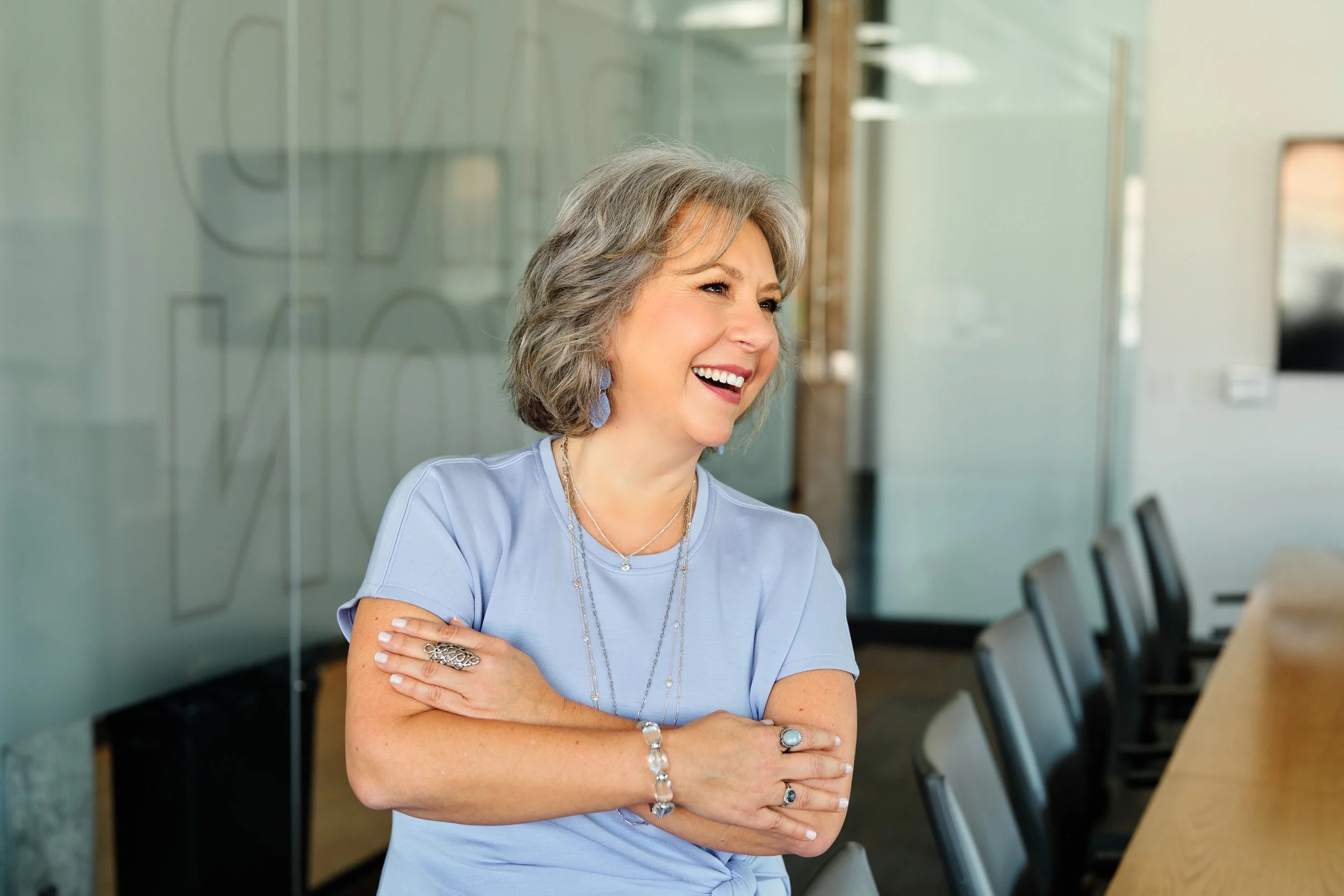 A woman with shoulder-length gray hair, wearing a light blue shirt, multiple jewelry pieces, and blue earrings, smiling and looking to her right in a modern office setting with glass walls and chairs.
