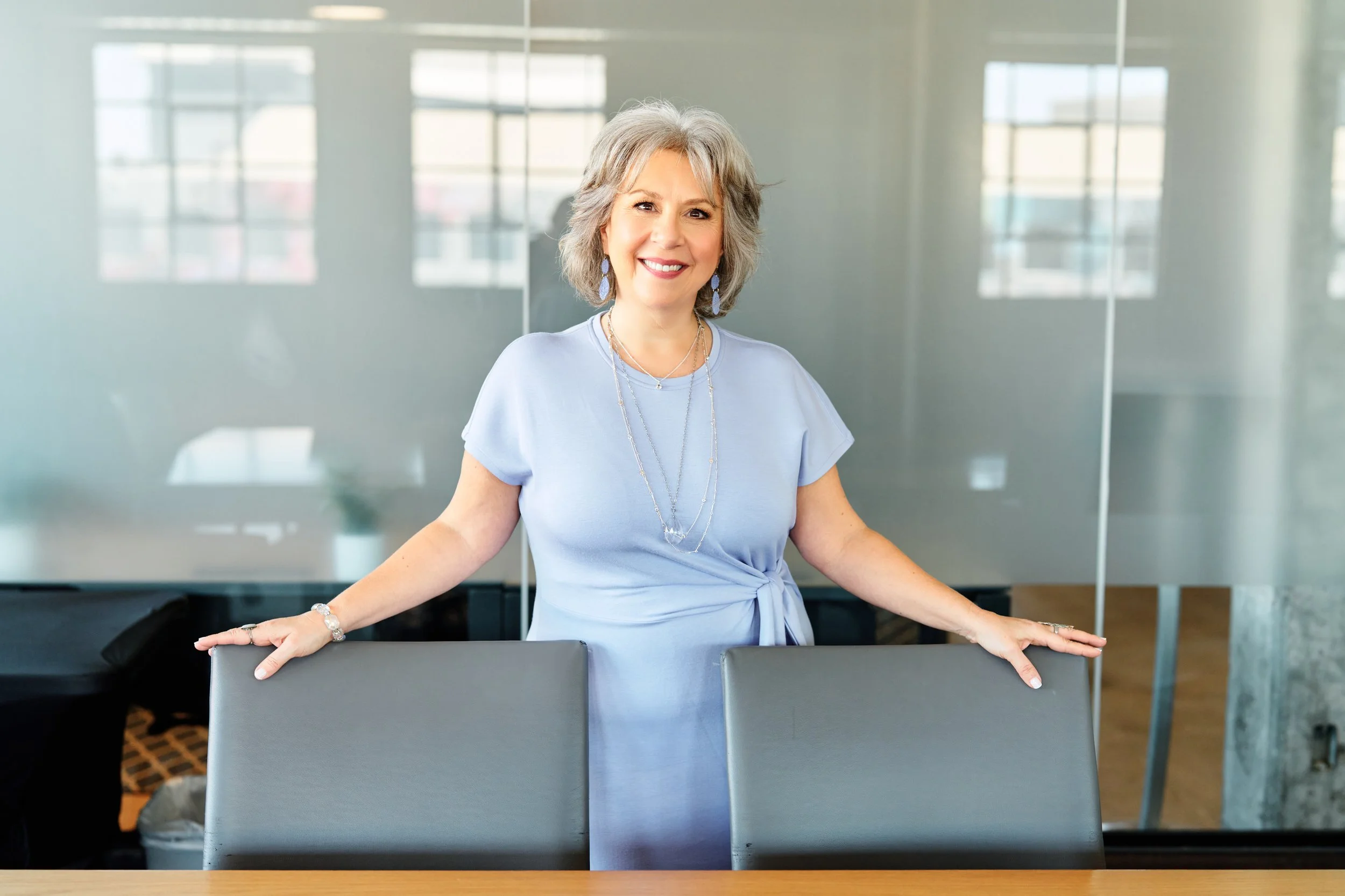 Smiling woman with gray hair wearing a light blue dress and jewelry, standing in a modern office conference room with glass walls and windows.