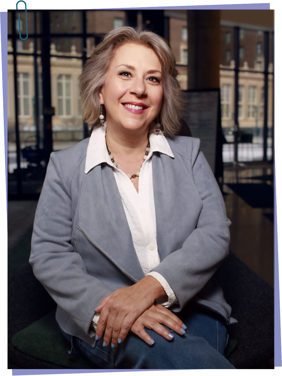 Portrait of a smiling middle-aged woman with short gray hair, wearing a light gray jacket over a white blouse, sitting indoors near large windows with a cityscape background.
