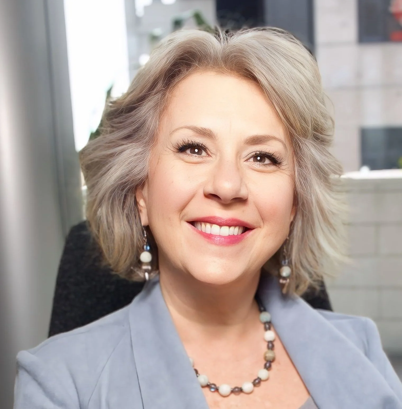 Close-up portrait of a smiling woman with shoulder-length gray hair, wearing earrings and a necklace, seated in an indoor setting with a window and blurred background.