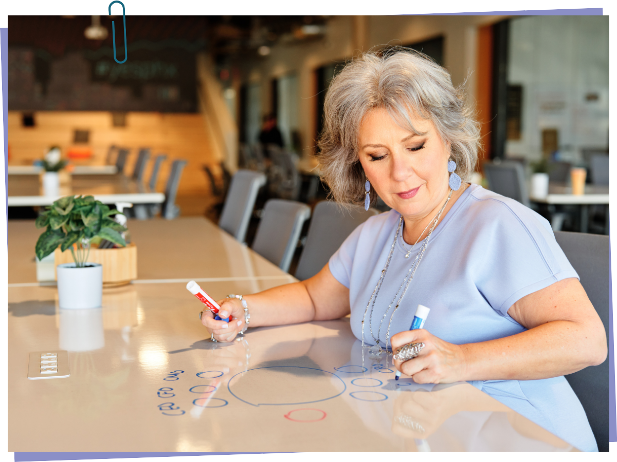 An older woman with gray hair and blue jewelry writing on a reflective white table in a brightly lit, modern office or cafe setting, with potted plants and chairs in the background.