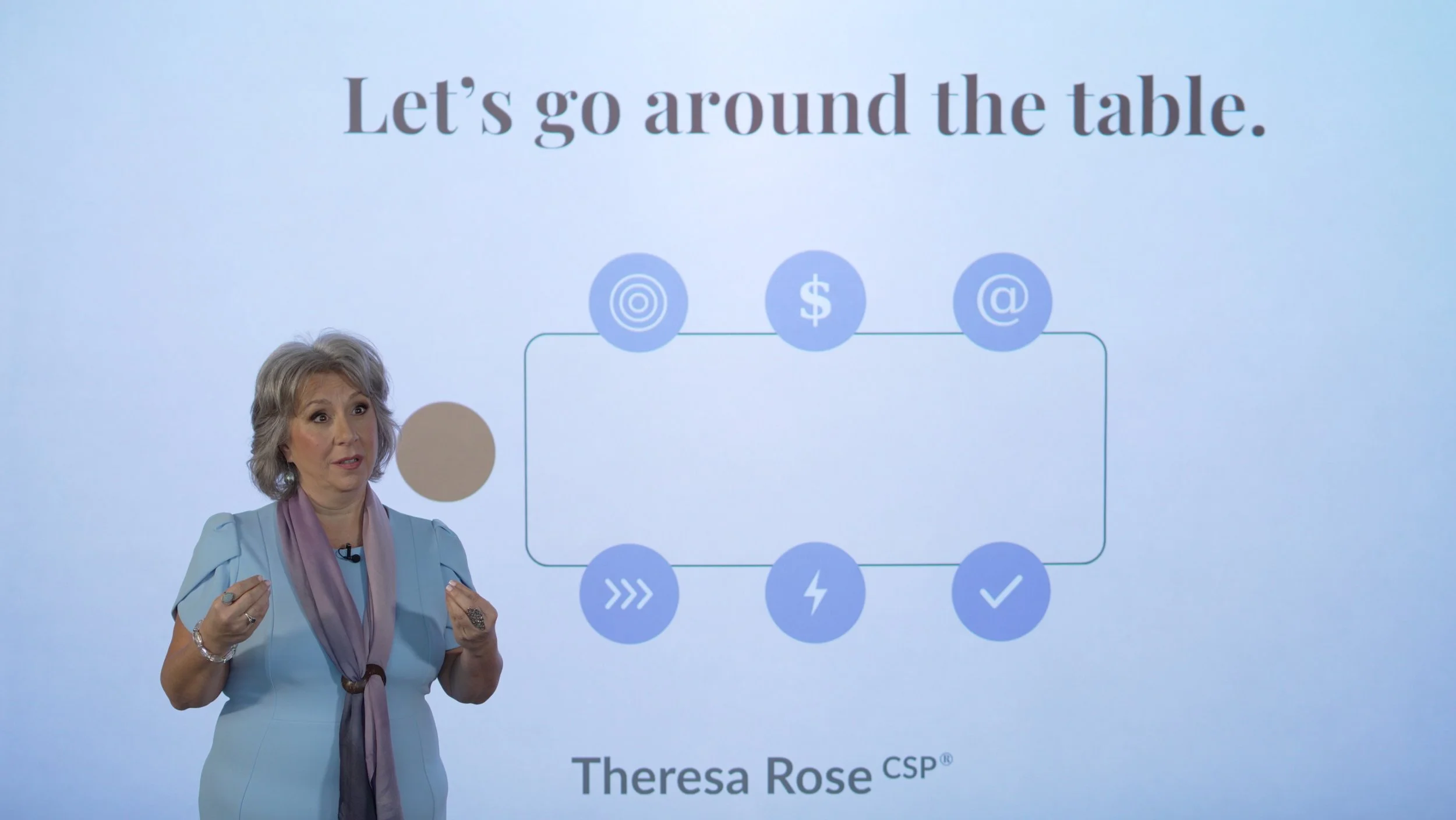 Theresa Rose CSP giving a presentation with a slide behind her that says 'Let's go around the table.' and shows icons of a target, dollar sign, at symbol, and other symbols. She has short gray hair, wears a light blue top, and a pink scarf.