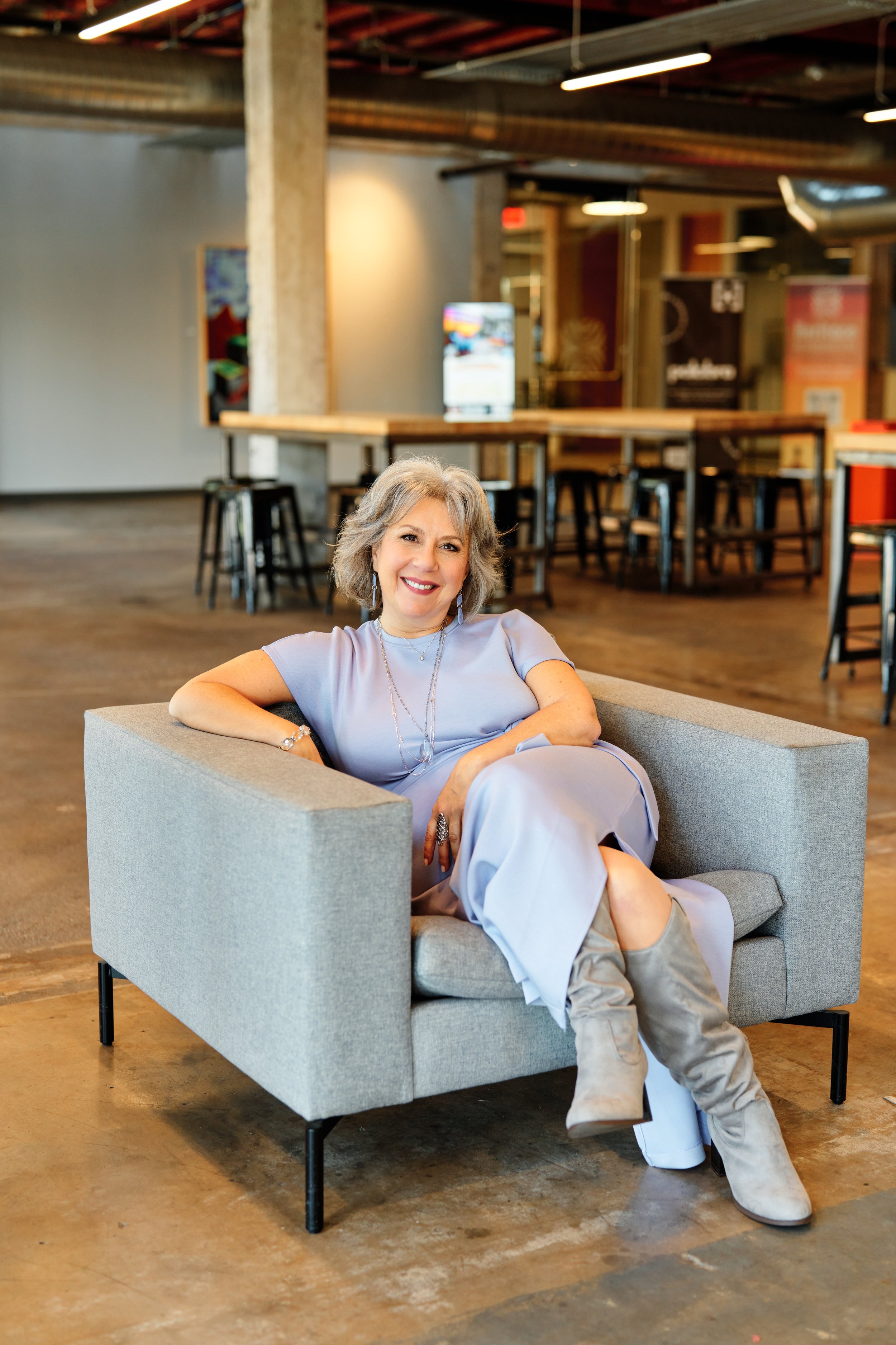 A woman with gray hair and a light purple dress is sitting on a gray sofa in an industrial-style space with exposed beams, concrete pillars, and wooden floors. She is smiling and relaxed, wearing beige boots and jewelry, with her arms resting on the sofa.