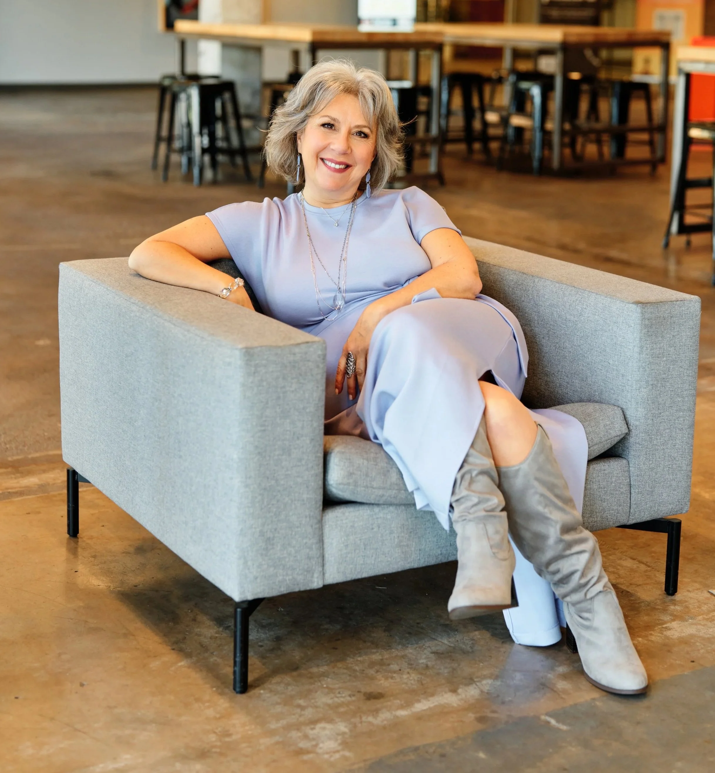 A middle-aged woman with gray, shoulder-length hair, smiling and sitting comfortably on a modern, light gray sofa with her arm resting on the armrest in a cozy, modern indoor space. She is wearing a light purple outfit, accessorized with long earrings, multiple necklaces, bracelets, and rings. The background features a wooden floor and tall tables with chairs, suggesting it might be a cafe or a community space.