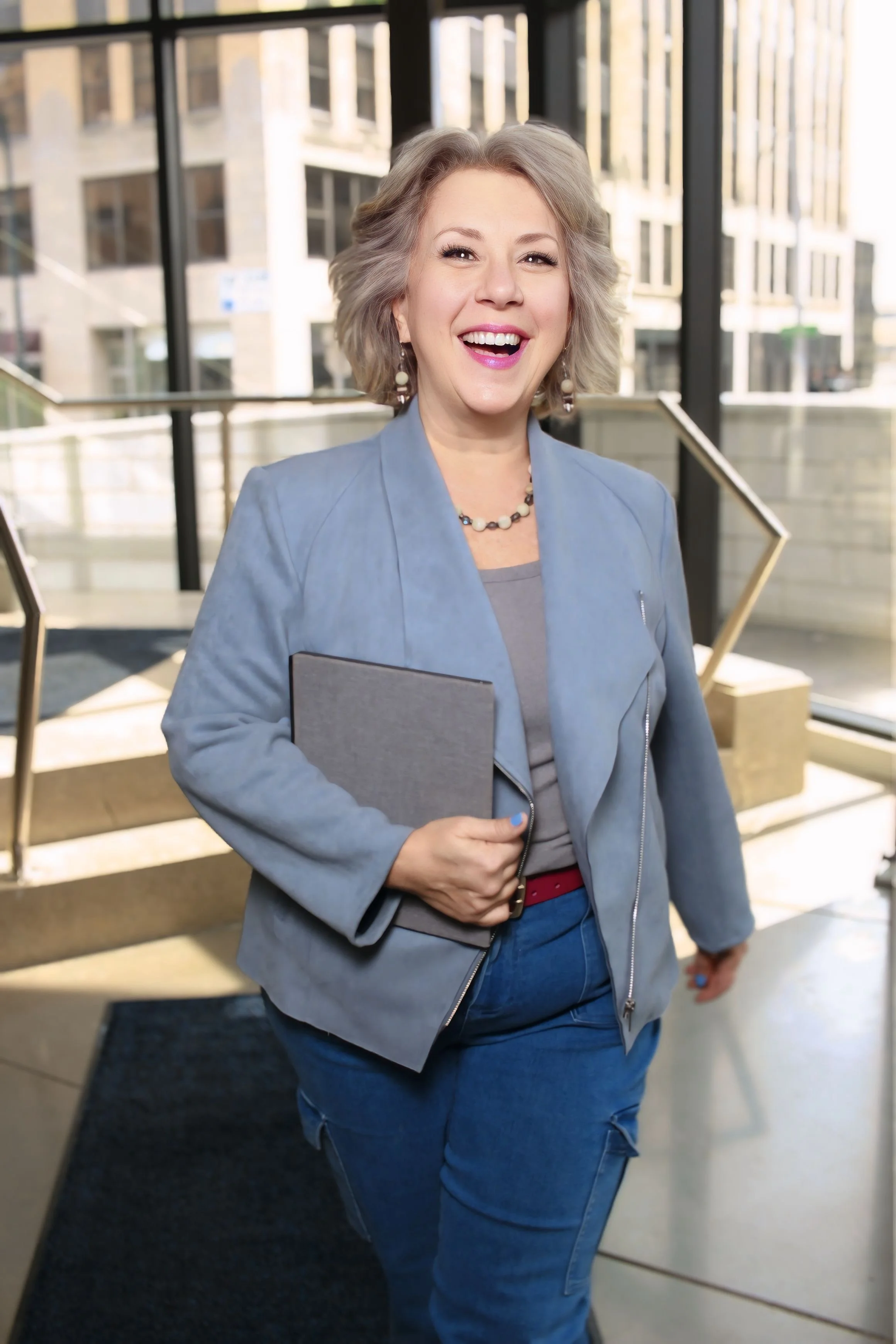 A woman with short gray hair smiling while holding a folder, wearing a blue blazer, a necklace, and earrings, standing inside a modern building with large glass windows and urban scenery in the background.