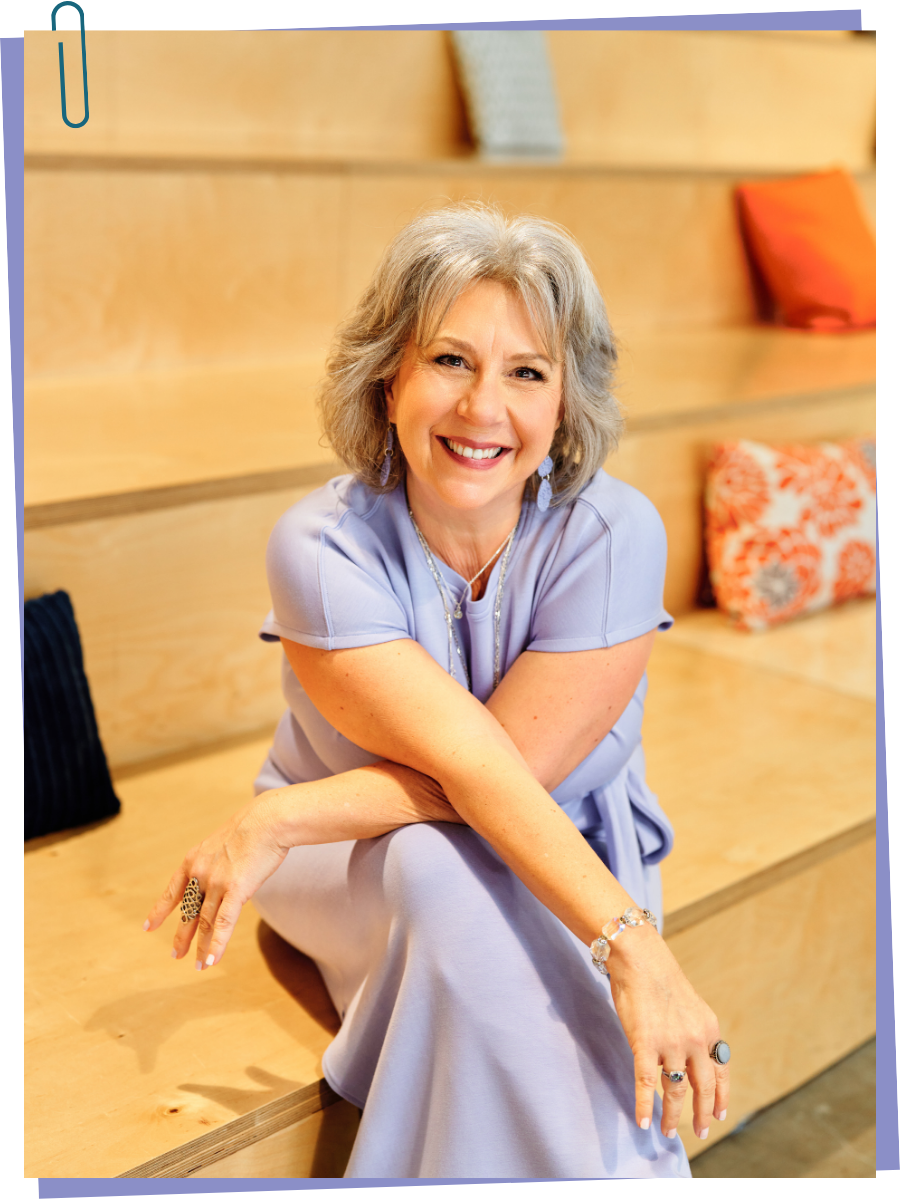 A smiling middle-aged woman with gray hair, wearing a light purple dress, sitting on wooden steps with colorful cushions in the background.