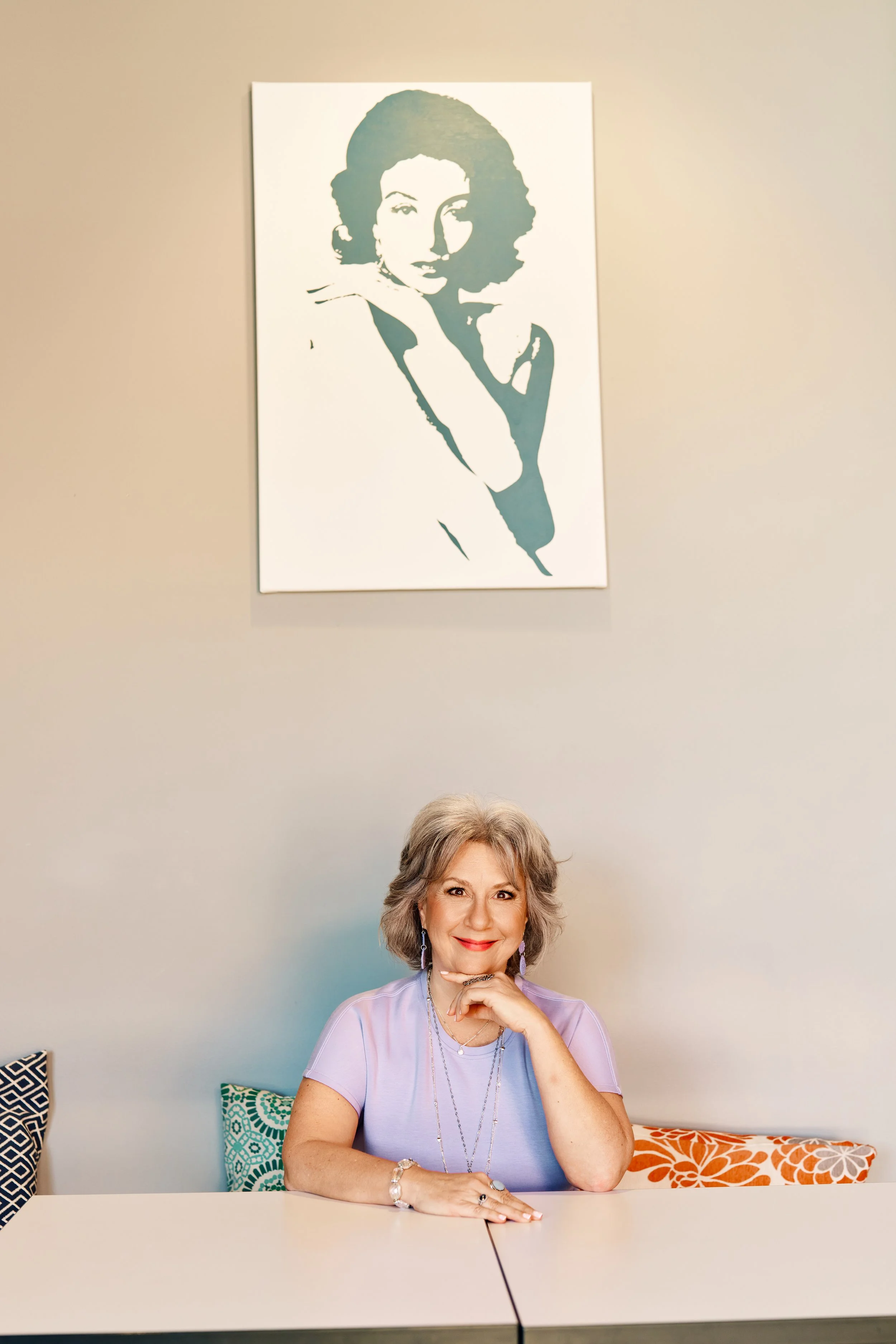 A woman with gray hair and a light purple top sitting at a white table in front of colorful cushions, with a portrait of a woman on the wall behind her.
