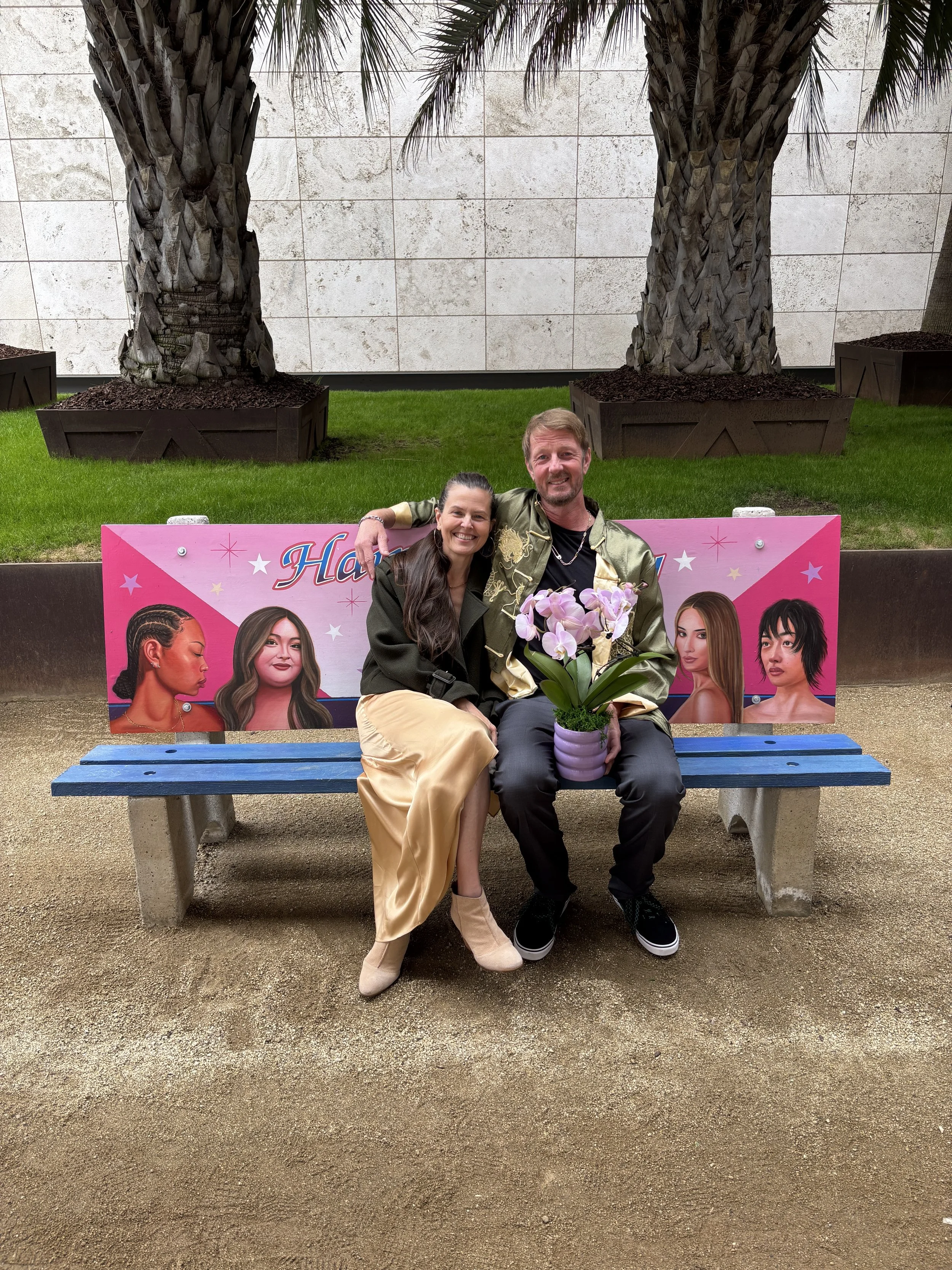 Tait Roelofs and Shelley De Soto seated on a bench by artist Alfonso Gonzalez Jr. at LACMA after the NEWH Oscar Brunch, holding an orchid centerpiece.