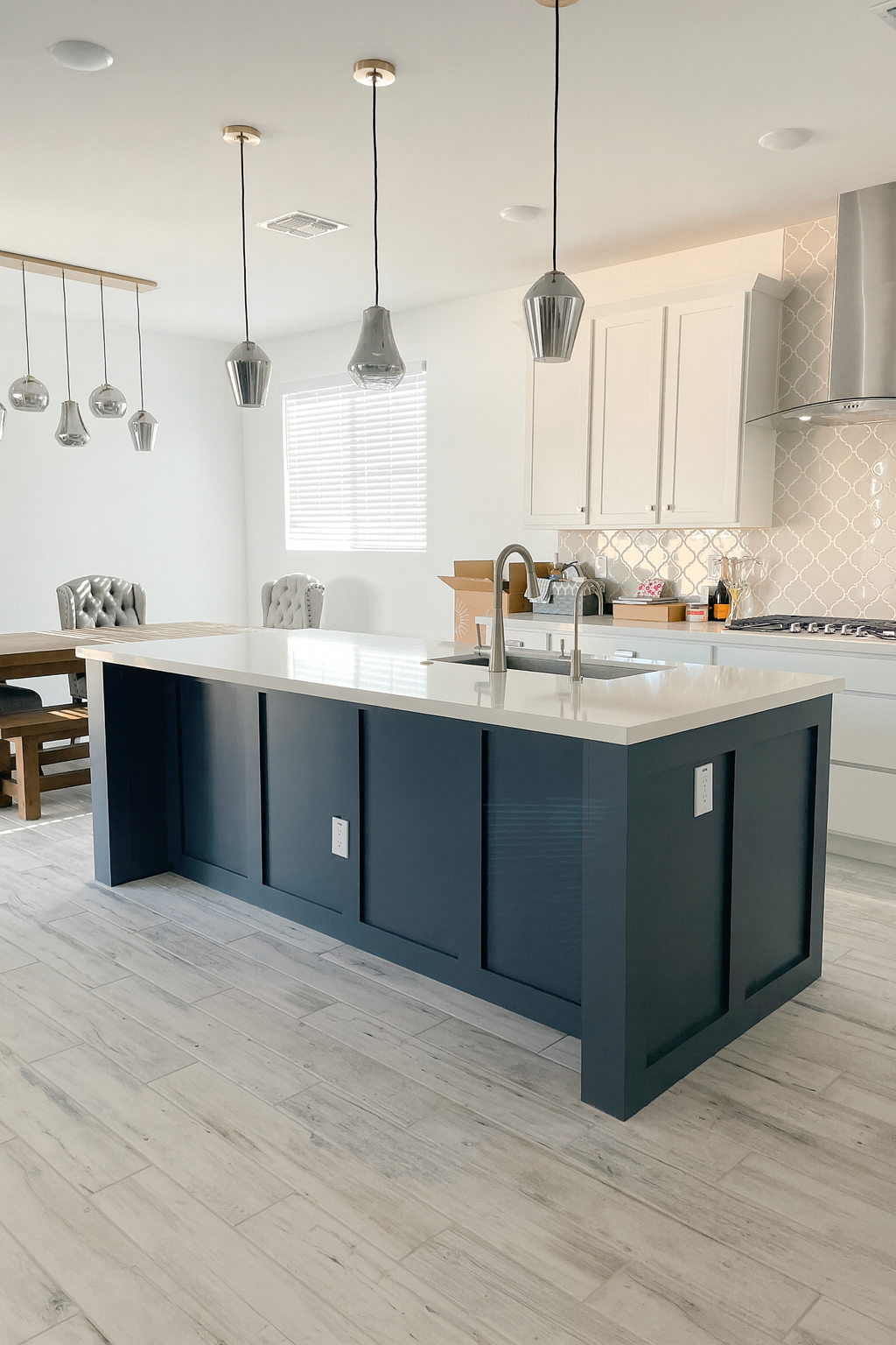 Modern kitchen with a navy blue island, white countertops, and white cabinets. Pendant lights hang above the island, and there is a window with blinds. Dining chairs are in the background.