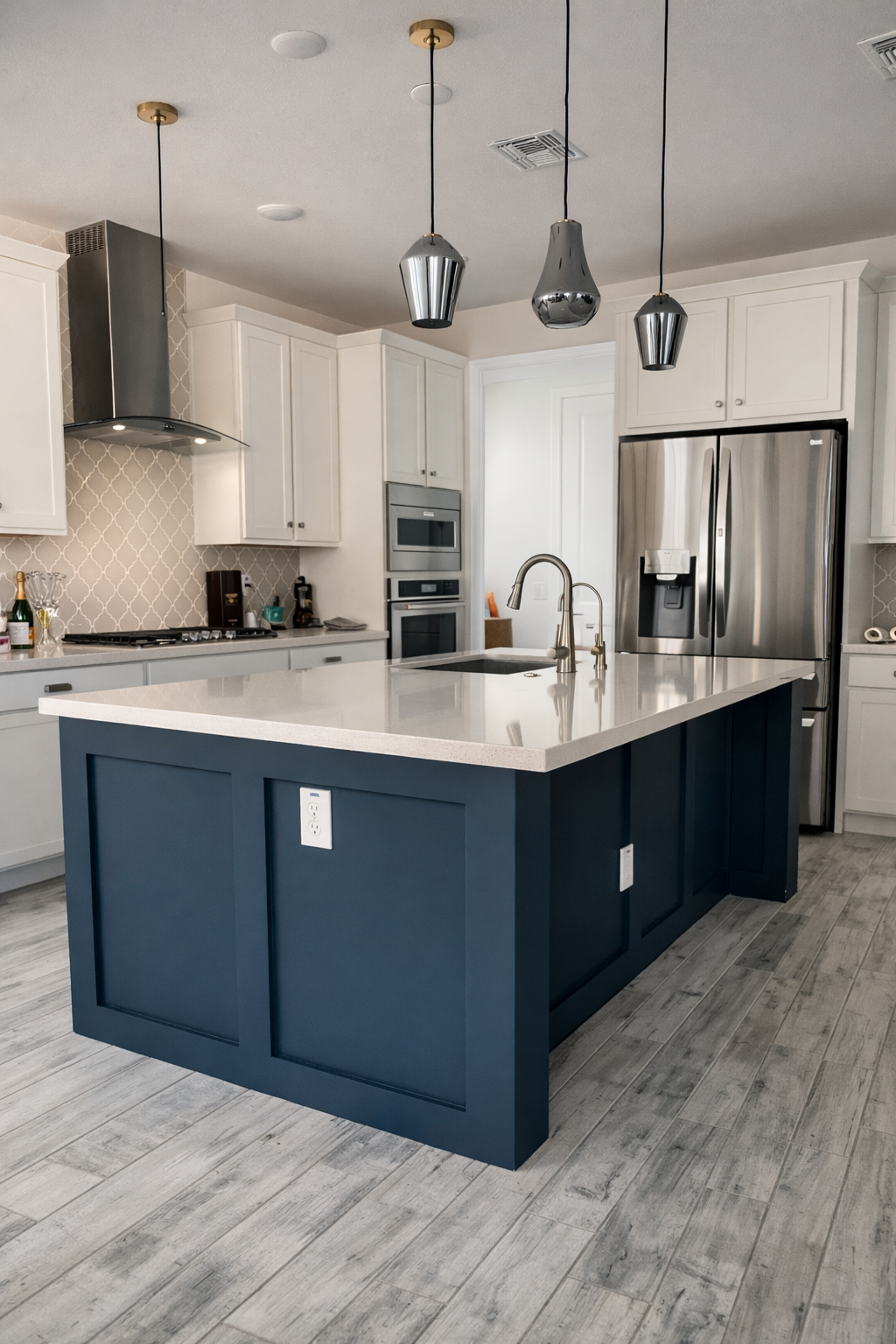 Modern kitchen with white cabinets, stainless steel refrigerator, built-in microwave and oven, and a blue island with a white countertop and a sink. The kitchen has three pendant lights hanging from the ceiling and light-colored wood flooring.