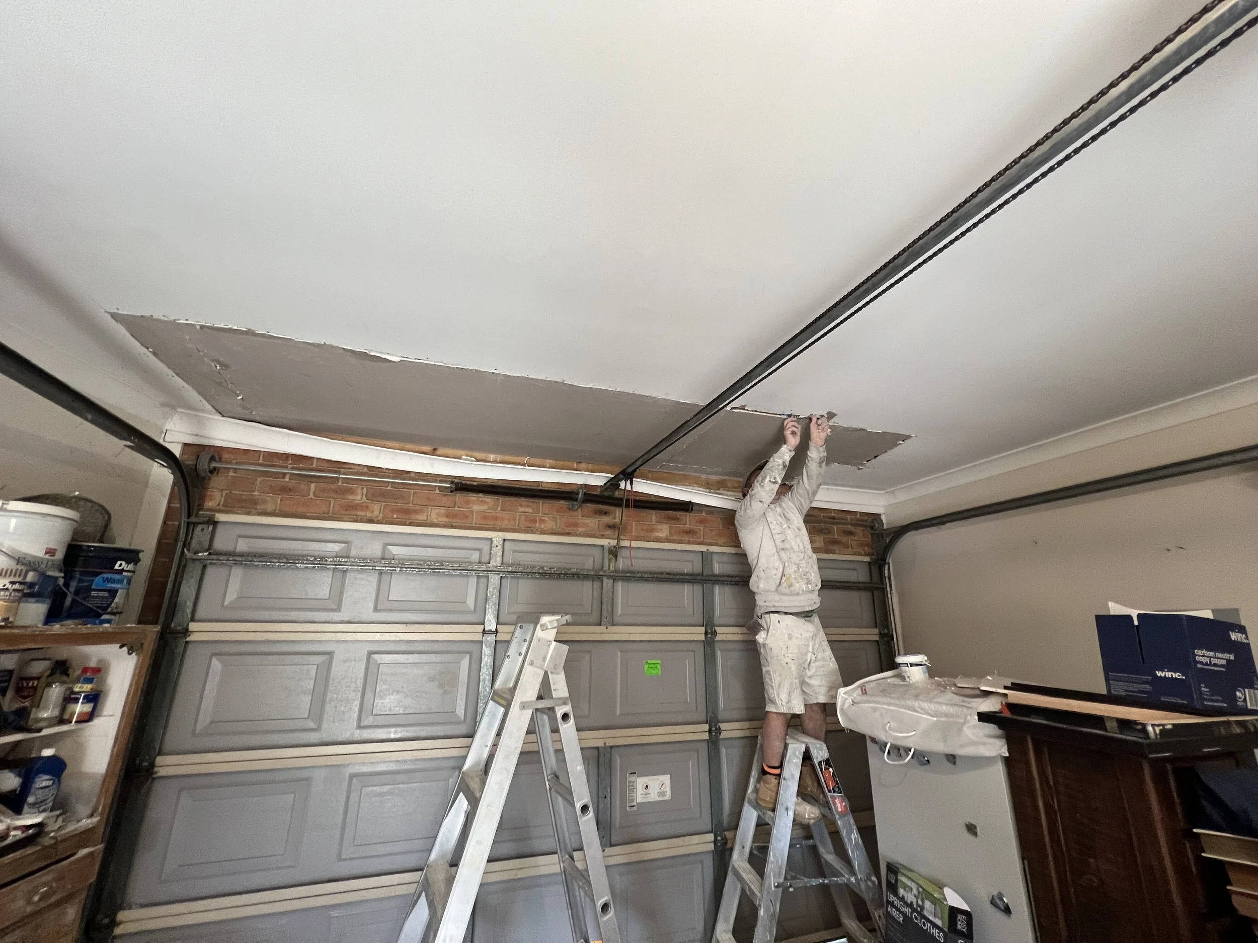 A person standing on a ladder working on a ceiling in a garage, with a partially installed ceiling panel and various supplies nearby.