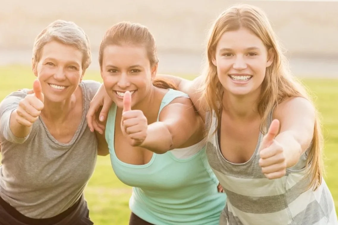 Three women smiling outdoors, wearing white tops, surrounded by green foliage.