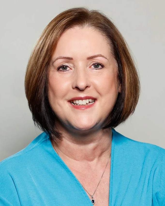 A woman with shoulder-length brown hair, smiling, wearing a blue top and a necklace, against a light gray background.
