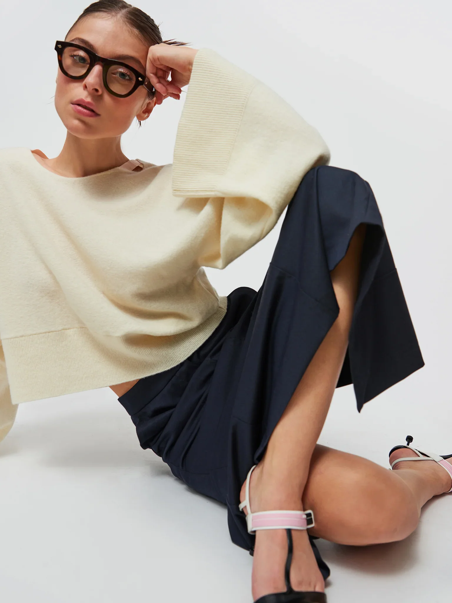 Une femme portant des lunettes noires, un sweat crème et une jupe noire, assise sur le sol blanc, portant une montre rose et une sandale blanche.