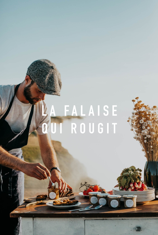 Homme en tenue décontractée et chapeau, préparant des confitures ou sauces sur une table en plein air avec la mer ou un océan en arrière-plan, et une composition florale dans un vase.