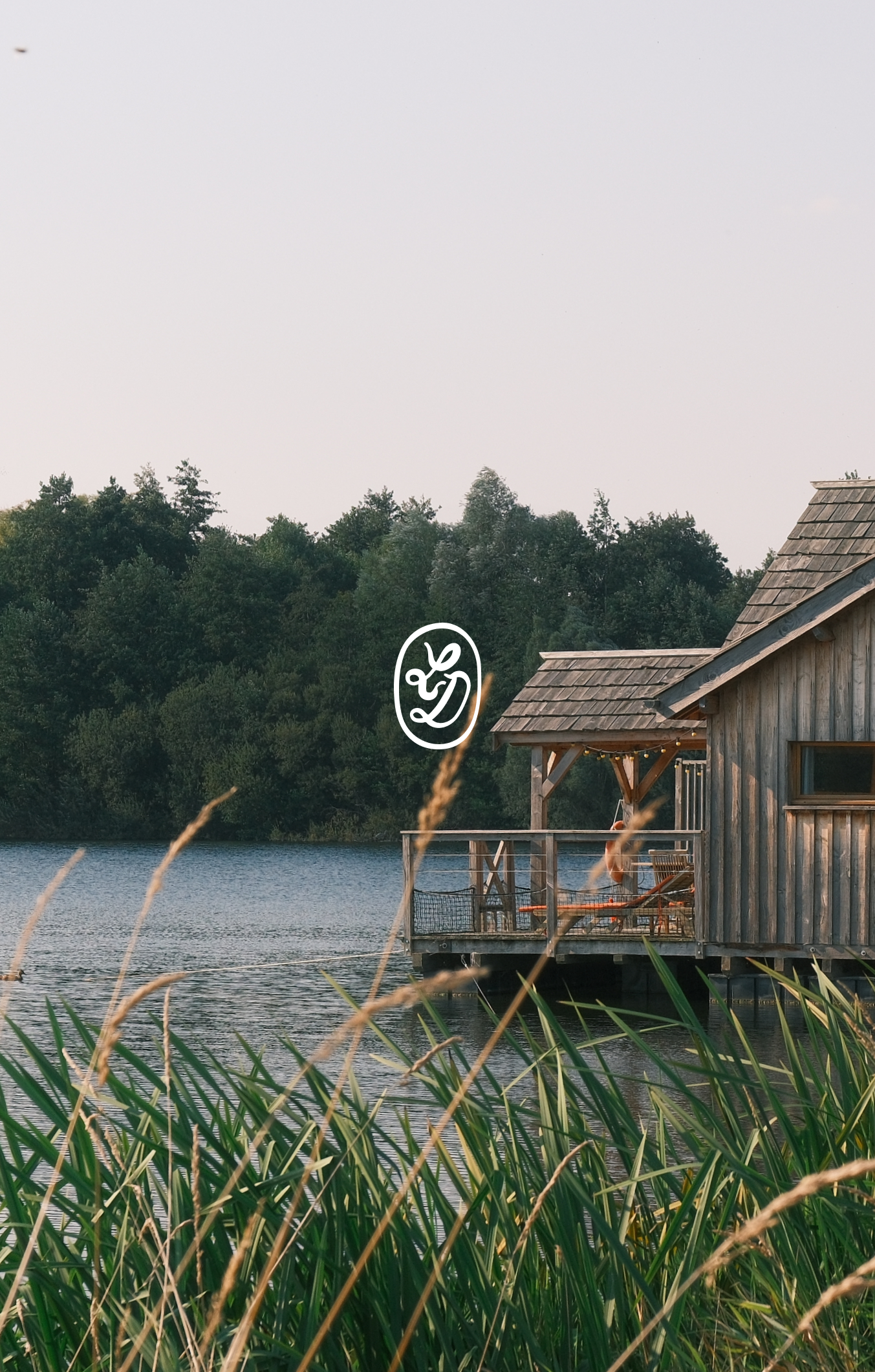 Une cabine en bois située au bord d'un lac, entourée de végétation, avec une terrasse en bois et un pont en bois. La scène est calme et naturelle, avec une forêt dense en arrière-plan.