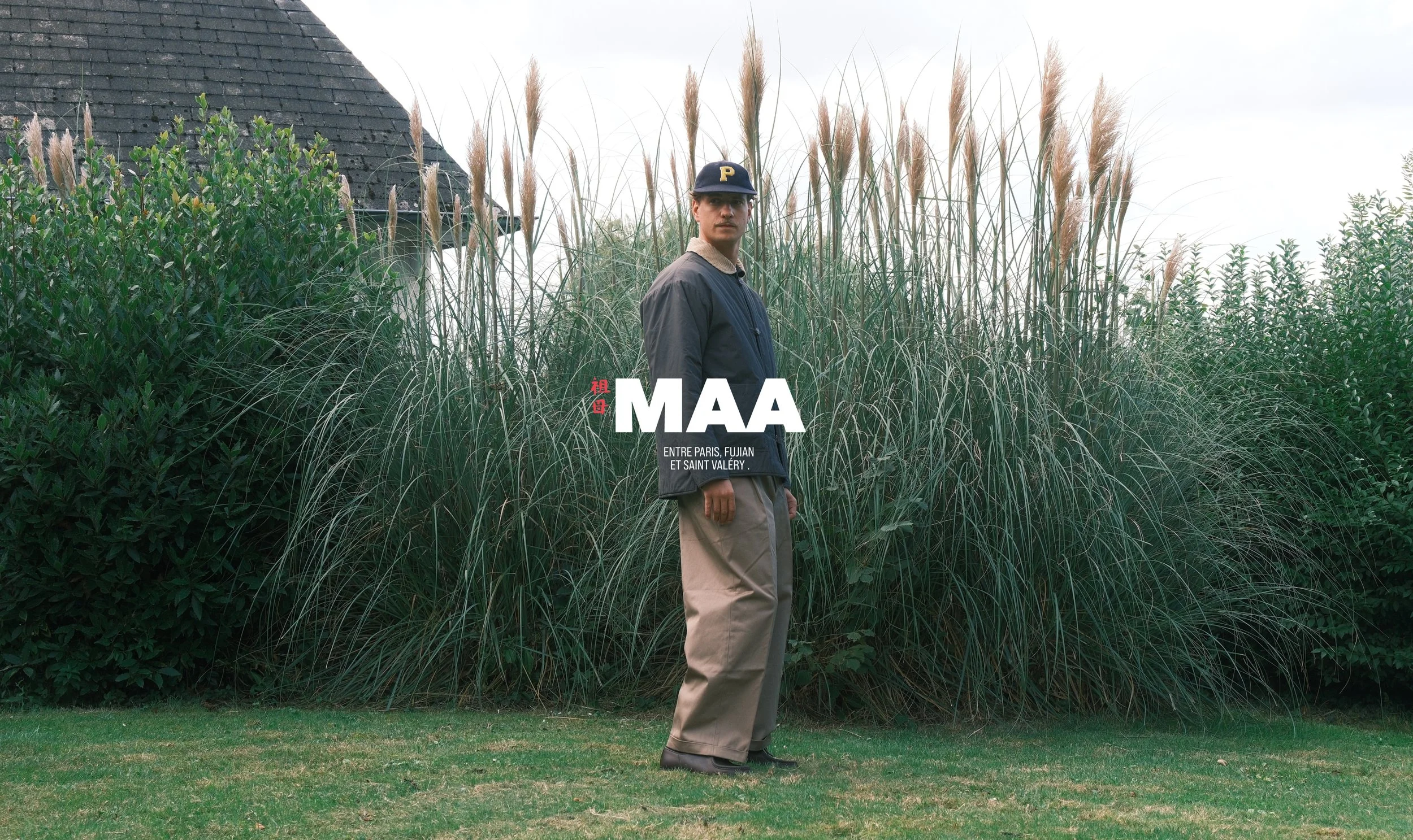 Jeune homme debout devant des hautes herbes, portant une casquette noire avec un P jaune, une veste noire et un pantalon beige, dans un environnement extérieur naturel avec une maison en arrière-plan