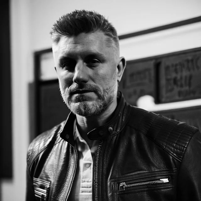 A black and white photograph of a man with short hair and a beard, wearing a leather jacket, standing indoors.