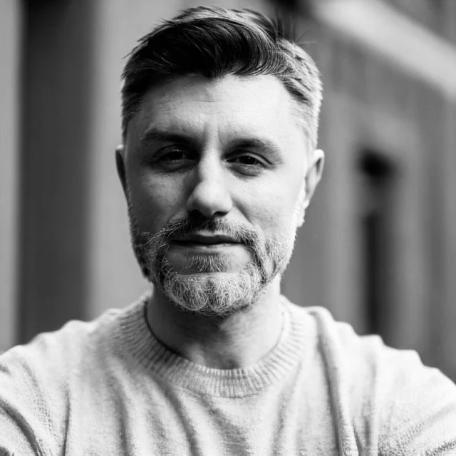 A black and white close-up portrait of a middle-aged man with a beard, smiling slightly, wearing a knit sweater, standing outdoors with a blurred background.