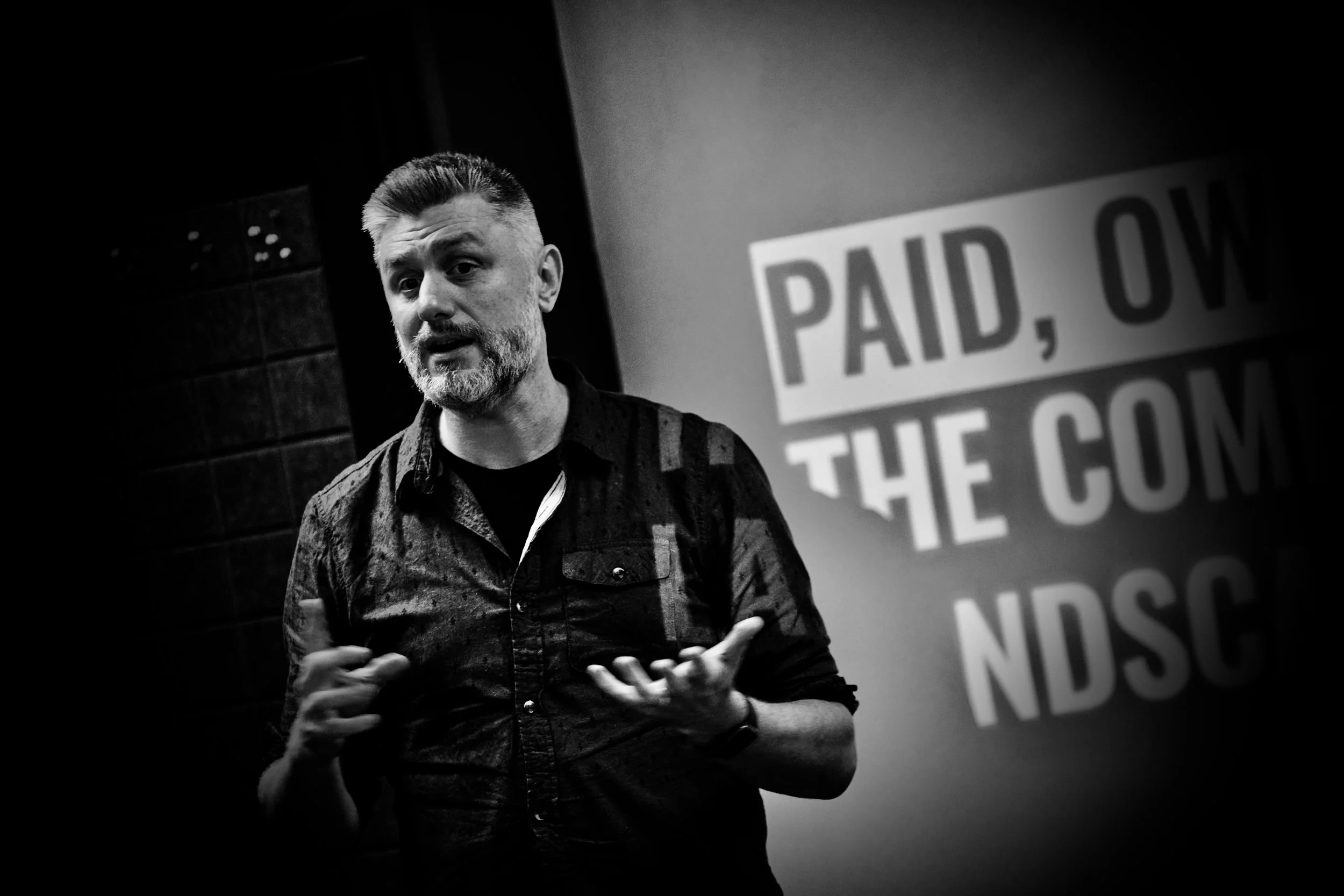 A man with a beard and short hair gesturing with his hands while speaking, standing in front of a sign that says 'PAID, OWE THE COM' in black and white.