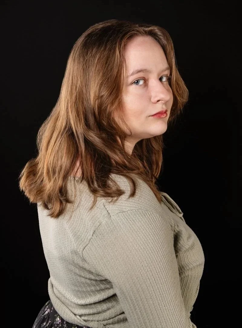 A woman with light brown, wavy hair and fair skin looking over her shoulder against a black background, wearing a light sweater.