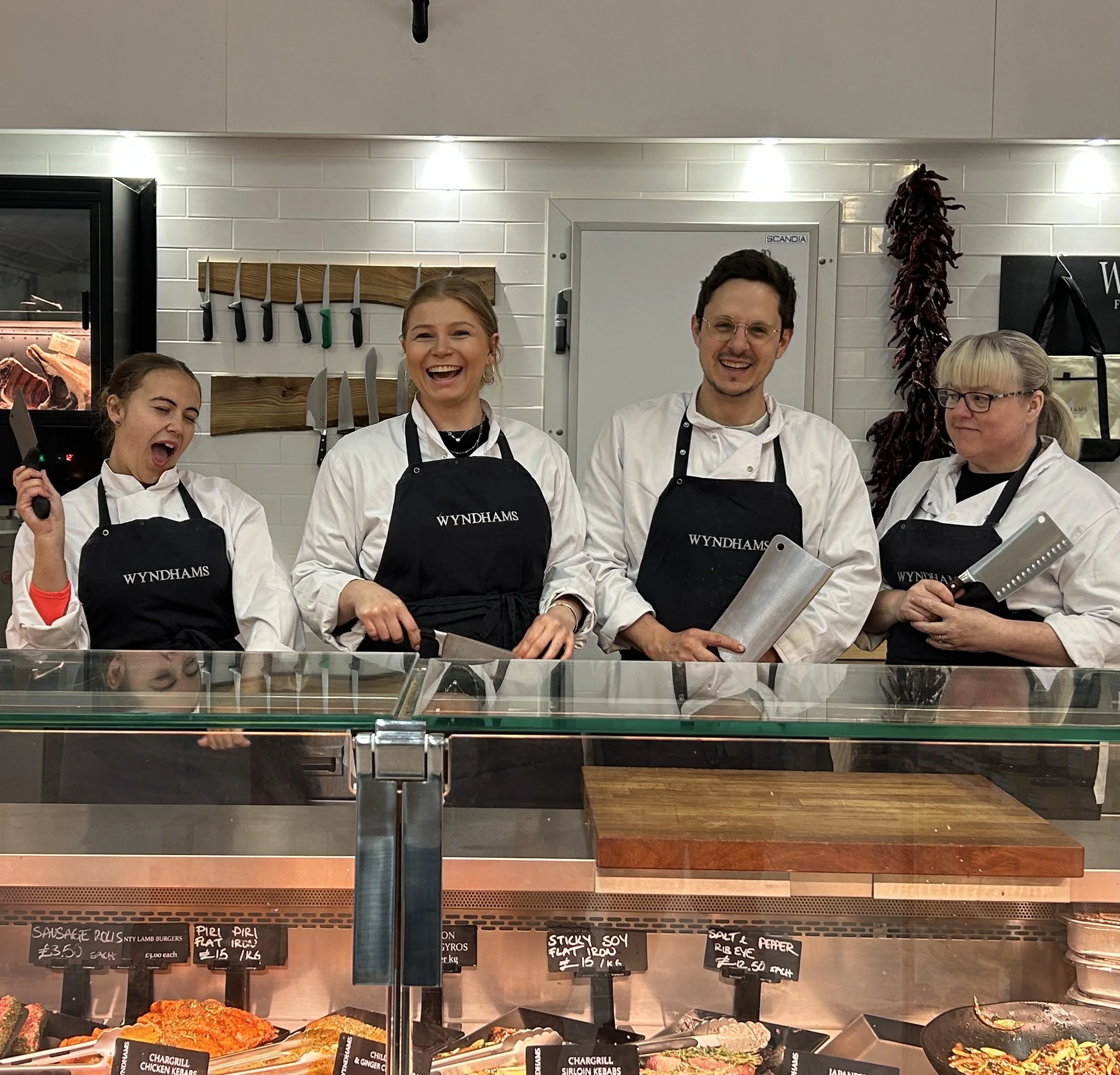Four chefs/ butchers wearing white uniforms and black aprons with 'WYNDHAMS' logo, smiling and holding knives or cleavers, standing behind a glass display case with meats and prepared foods in a modern kitchen or deli environment.
