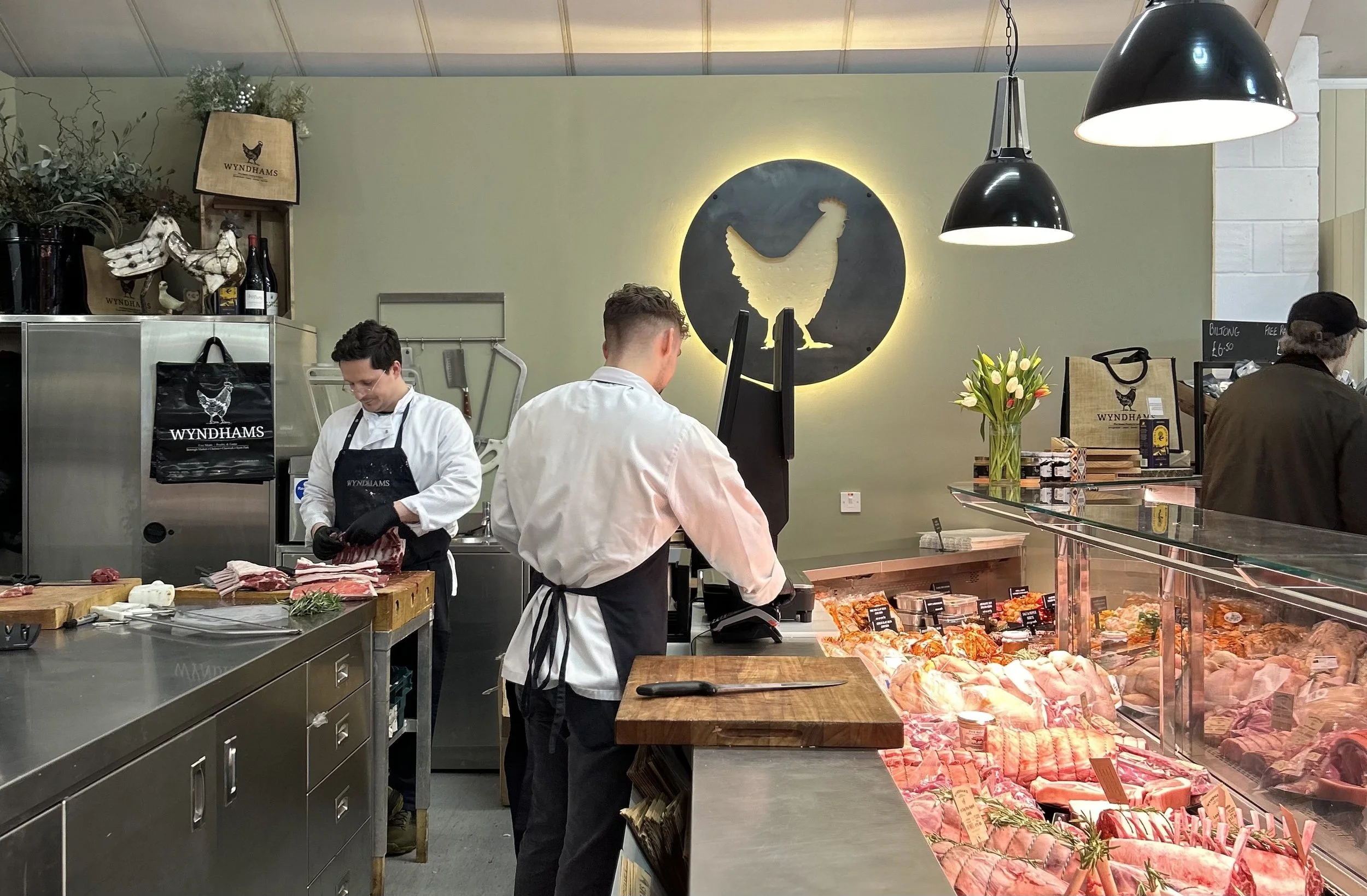 Butchers working behind a glass display case filled with various cuts of meat inside a restaurant or butcher shop. One butcher is slicing meat, and another is operating a scale. The interior features a black circular sign with a hen silhouette and yellow backlight, pendant lights, and decorative items like flowers and bags.