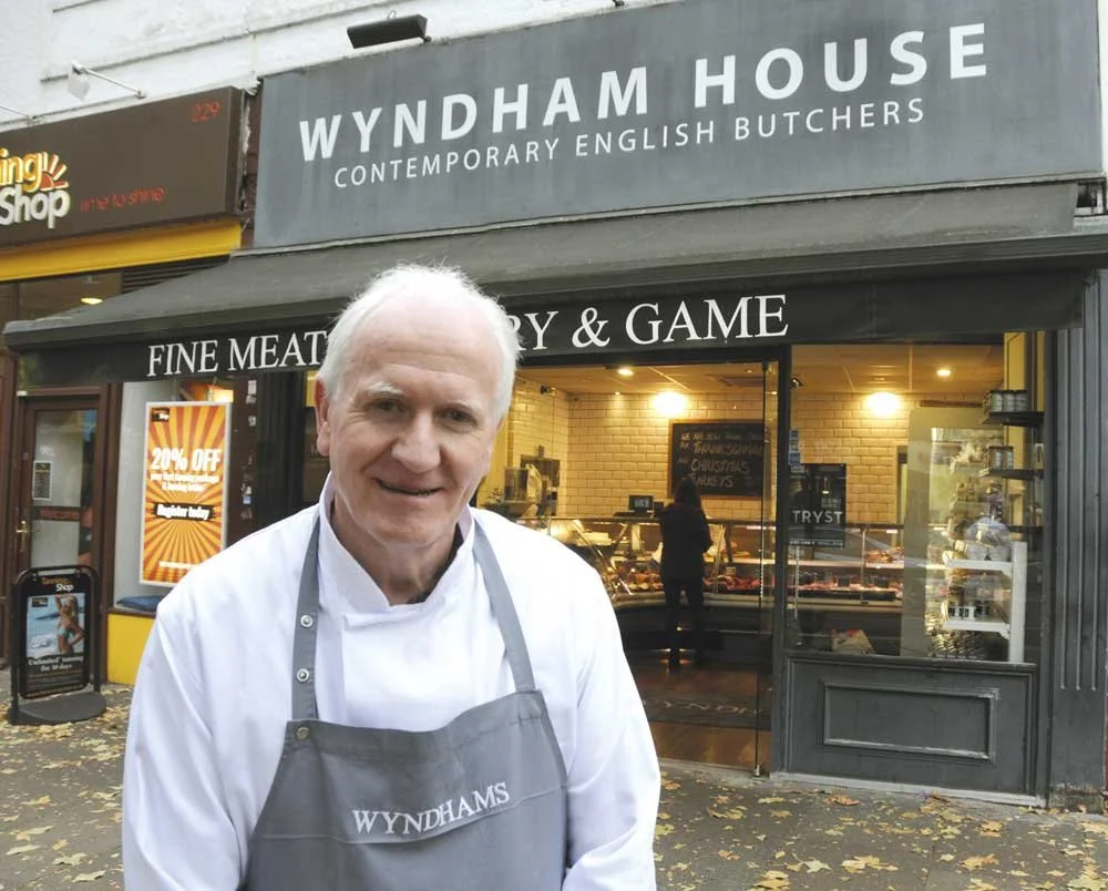 A man in a white chef's jacket and gray apron standing outside Wyndham House, a contemporary English butchers shop, with a person inside visible through the window.