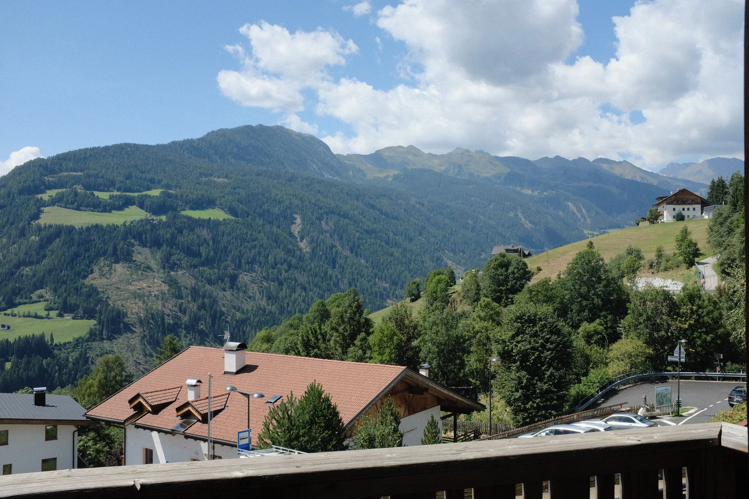 Häuser mit roten Dächern vor grünen Hügeln und Bergen, bewölkter Himmel