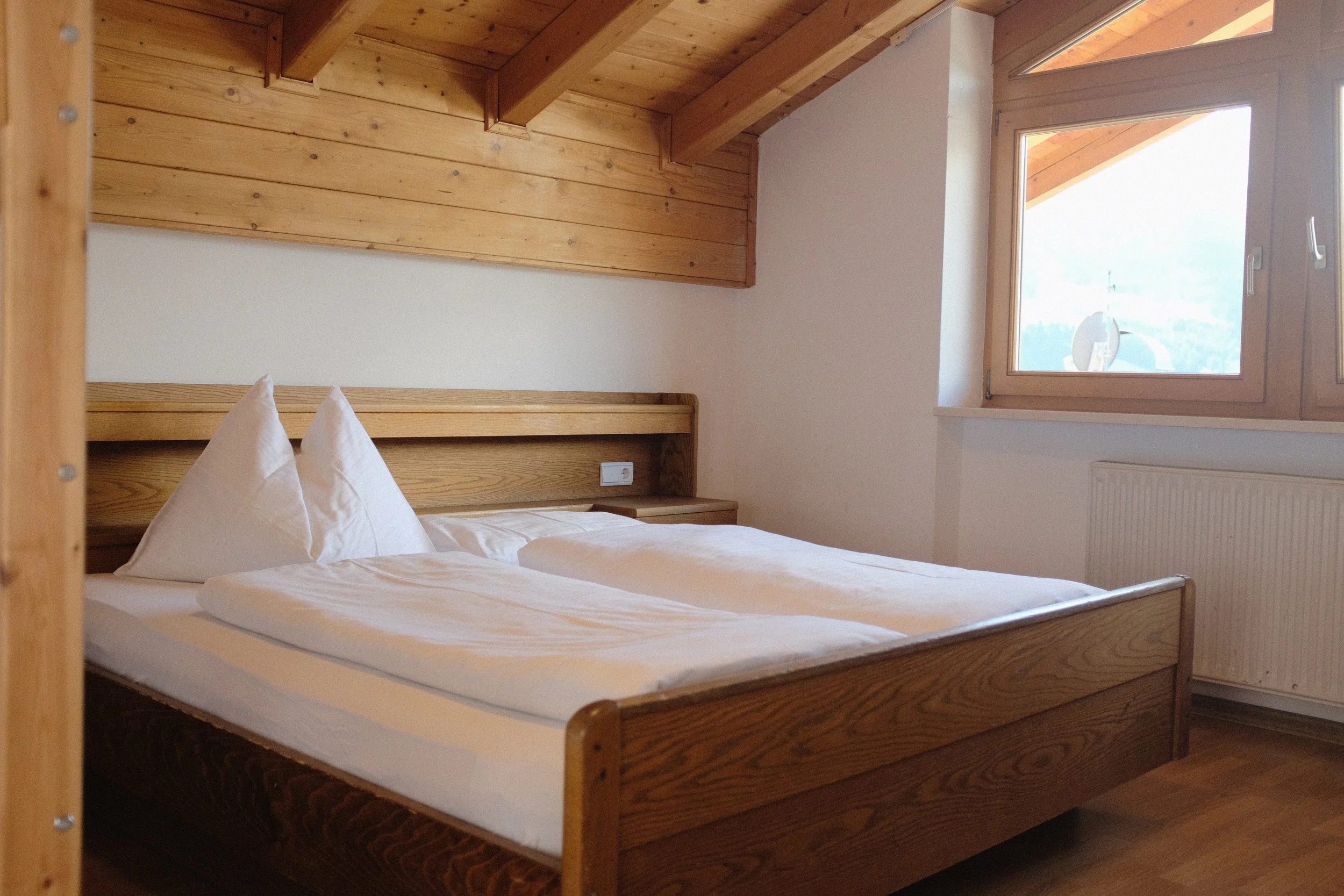 Bett in einem Schlafzimmer mit Holzwand und -decke, zwei weiße Kissen, Fenster mit Ausblick auf einen Himmel und Bäume.