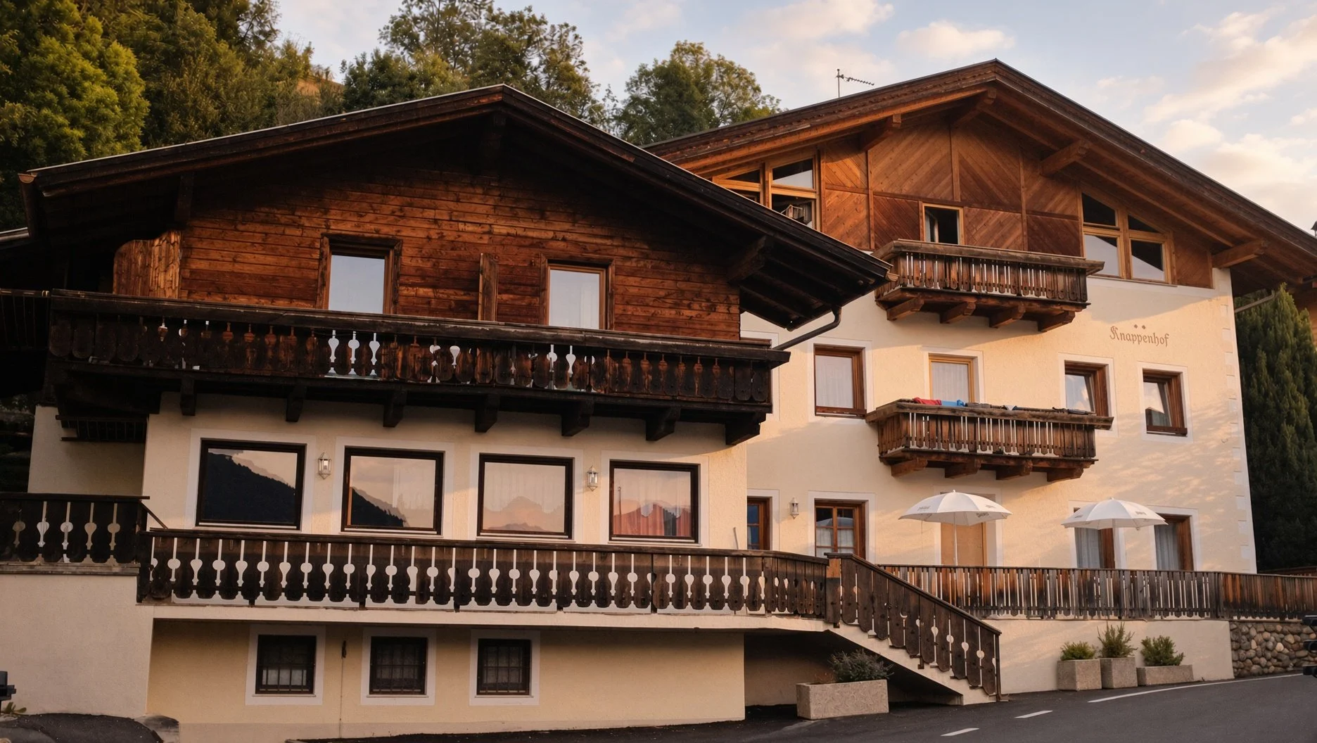 Mehrstöckiges Chalet mit Holzbalkonen, weißen Wänden und einem Berg im Hintergrund bei Sonnenuntergang.