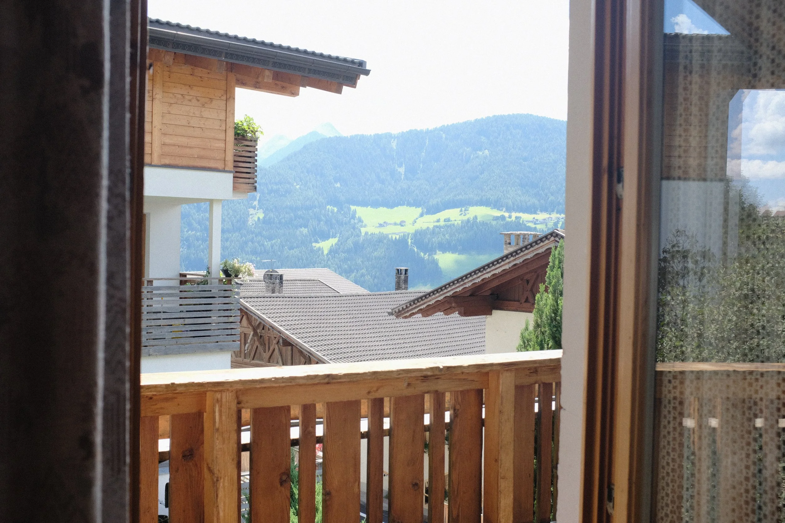 Blick aus einem Fenster auf einen Balkon mit Holzzäunen, darunter Dächer anderer Häuser und im Hintergrund grüne Hügel und Berge bei sonnigem Himmel.
