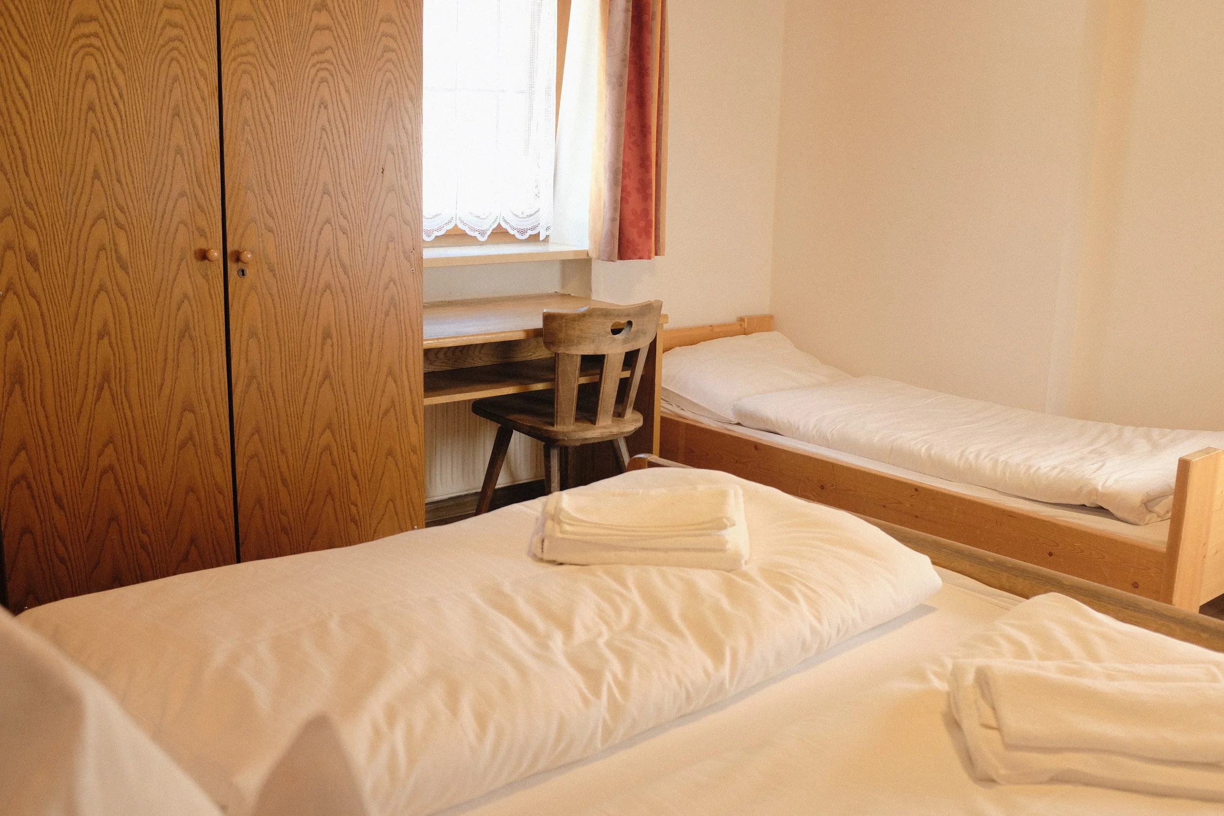 Ein kleines Schlafzimmer mit zwei Betten, einem Holztisch und Stühlen, einem Kleiderschrank und einem Fenster mit weißen Vorhängen und roten Gardinen.