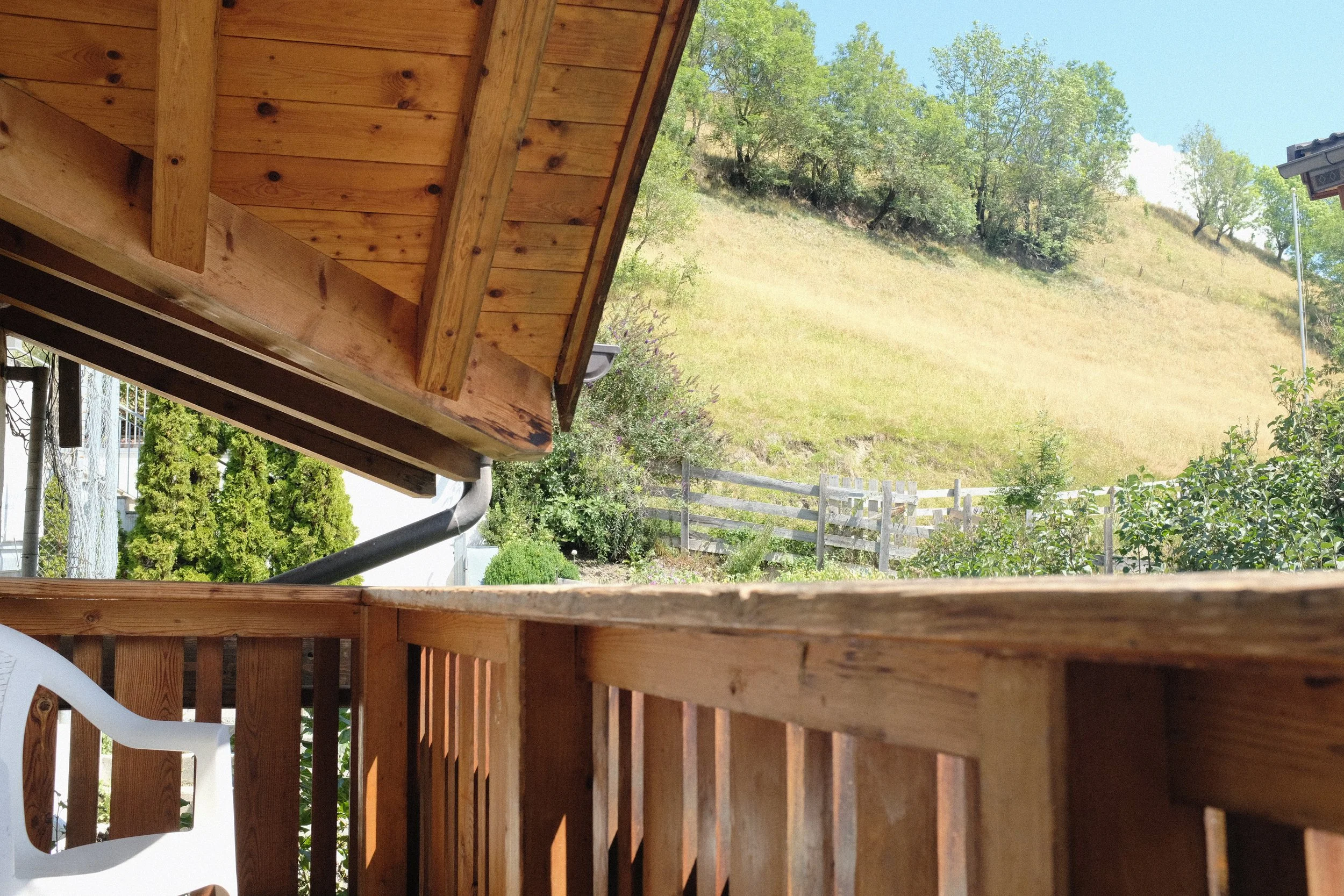 Blick vom Balkon eines Holzhauses auf einen grünen Hang mit Bäumen, einen Zaun und den blauen Himmel.