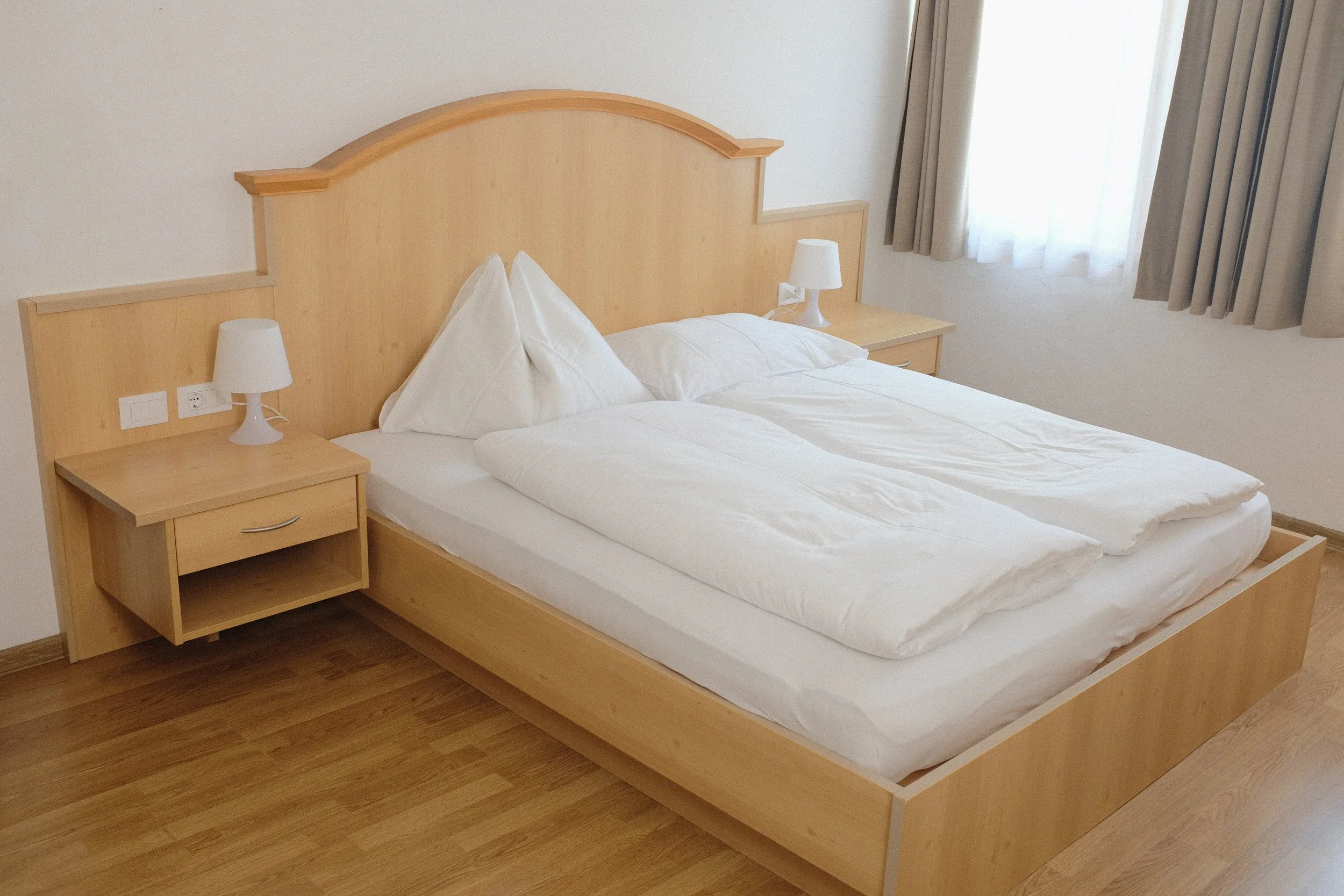 Ein Bett mit weißer Bettwäsche und zwei weißen Kissen in einem schlichten Hotelzimmer, mit einem Holz-Nachtisch und Tischlampen auf beiden Seiten.
