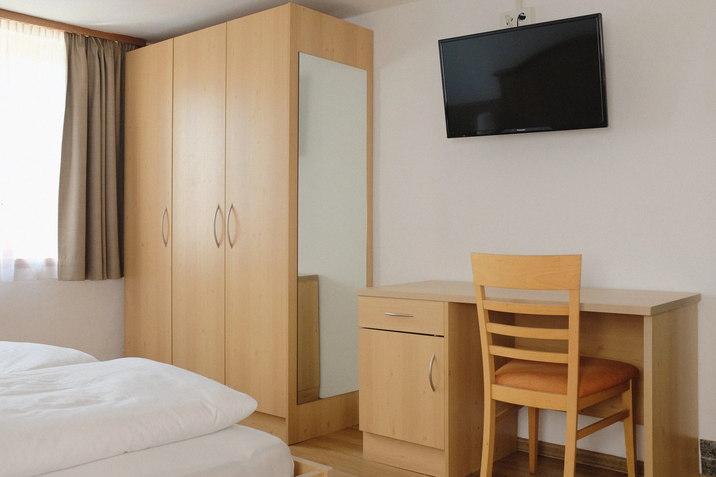 Ein Schlafzimmer mit einem Bett, einem Kleiderschrank, einem Wandspiegel, einem kleinen Schreibtisch und einem Stuhl, sowie einem Fernseher an der Wand.