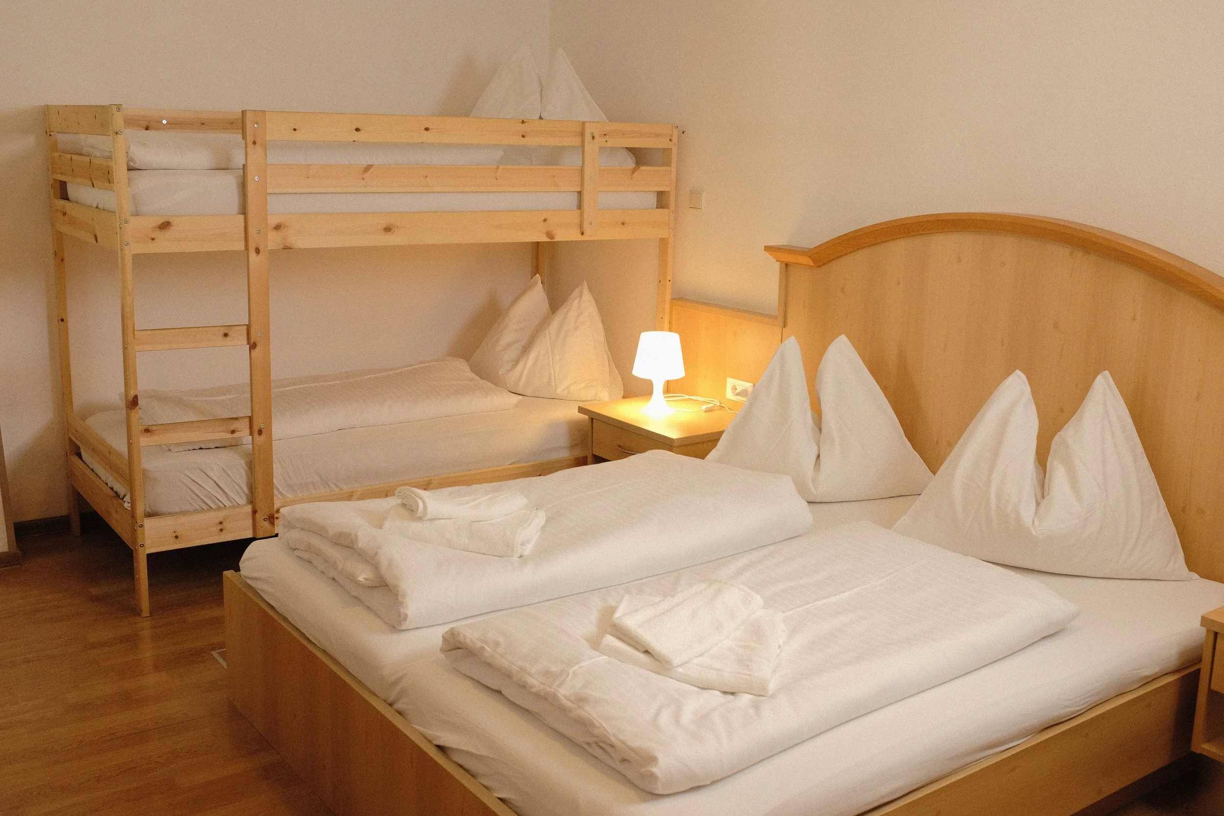 Ein Hotelzimmer mit zwei Betten, Schlafsack und Kissen. Das obere Bett ist ein improvisiertes Etagenbett aus Holz. Es gibt eine Nachttischlampe und eine Wandsteckdose.