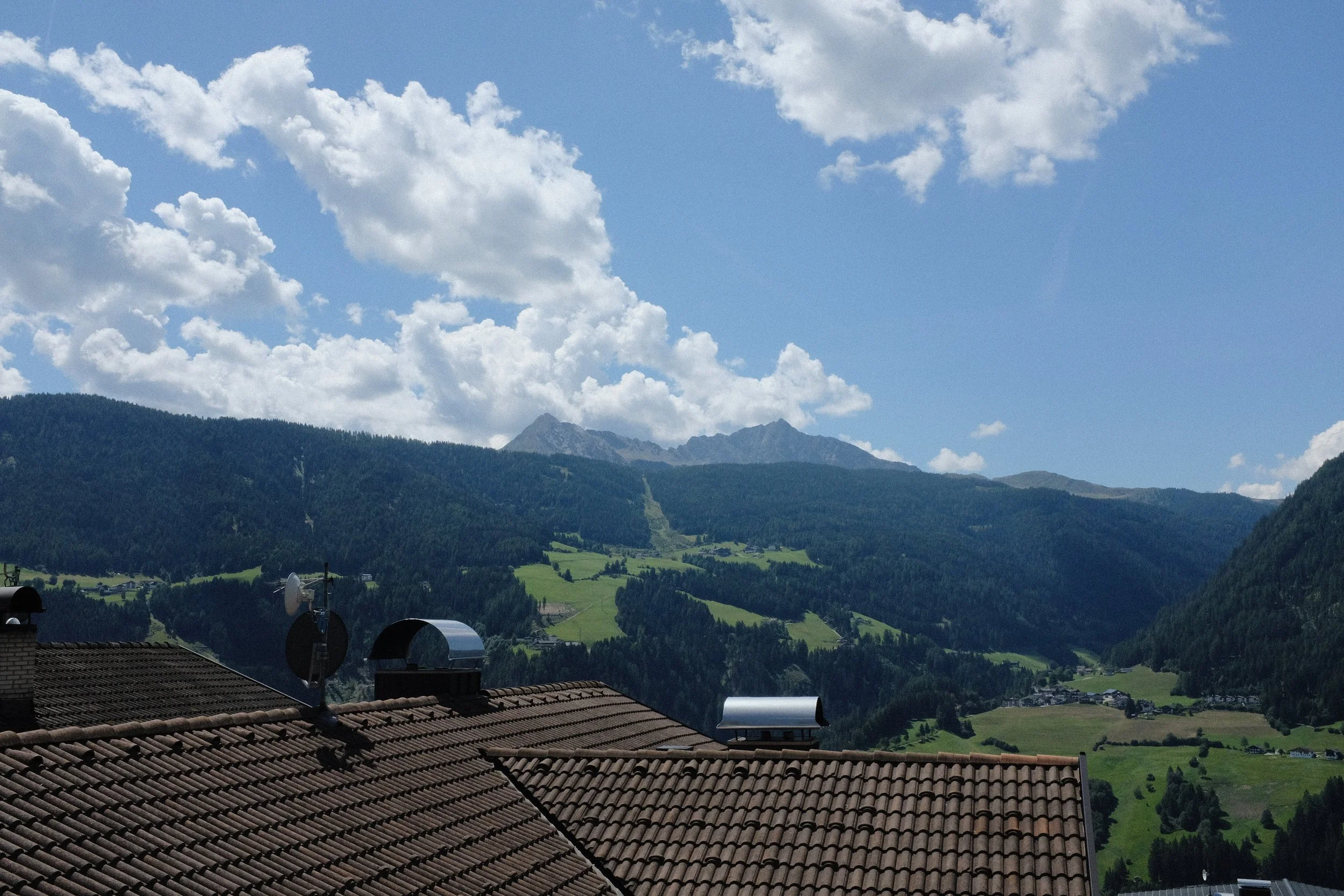 Berglandschaft mit Wald, grünen Wiesen und einem blauen Himmel mit Wolken, im Vordergrund Dachzimmer mit Antennen.