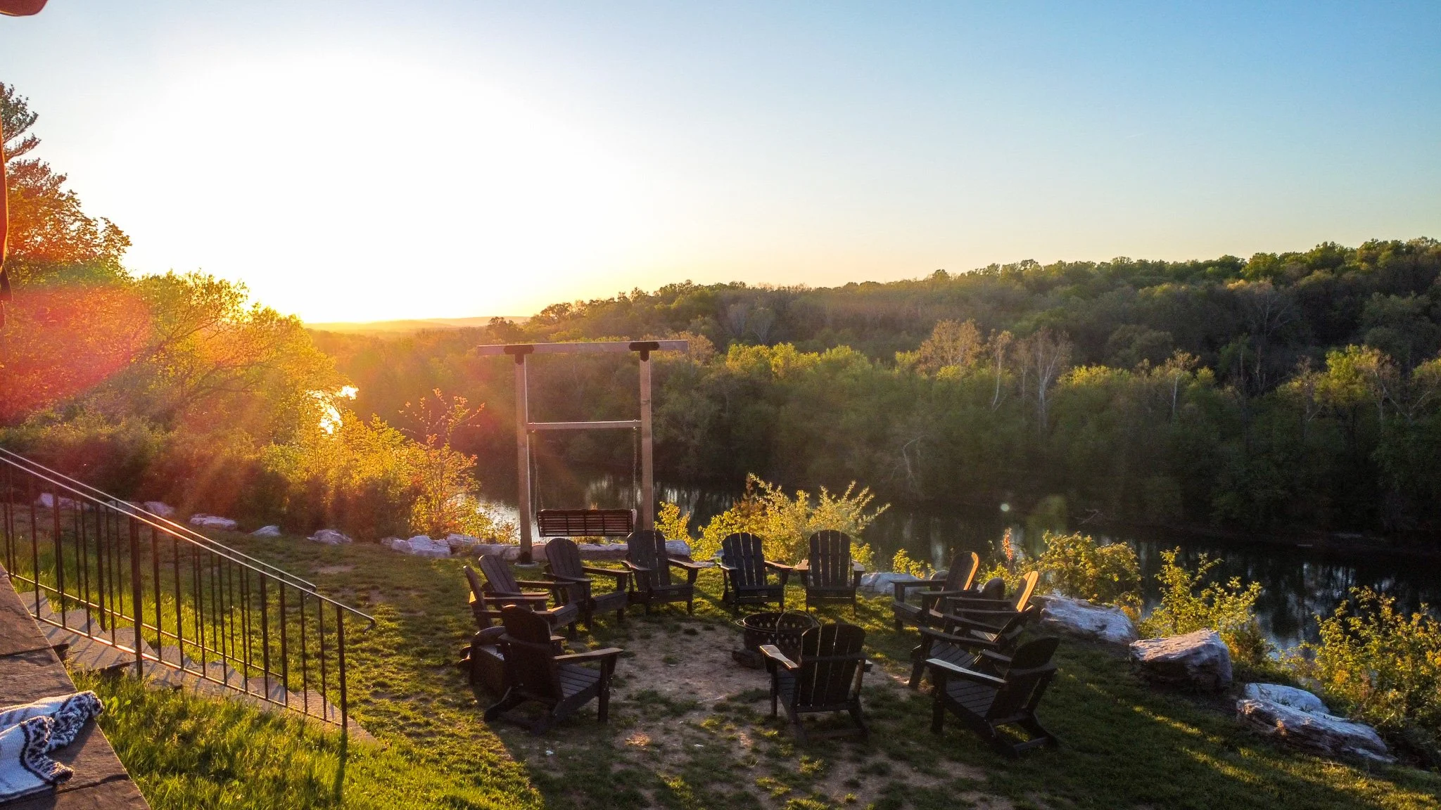 Sunset view, wooden chairs around a fire pit, overlooking a river and forest.