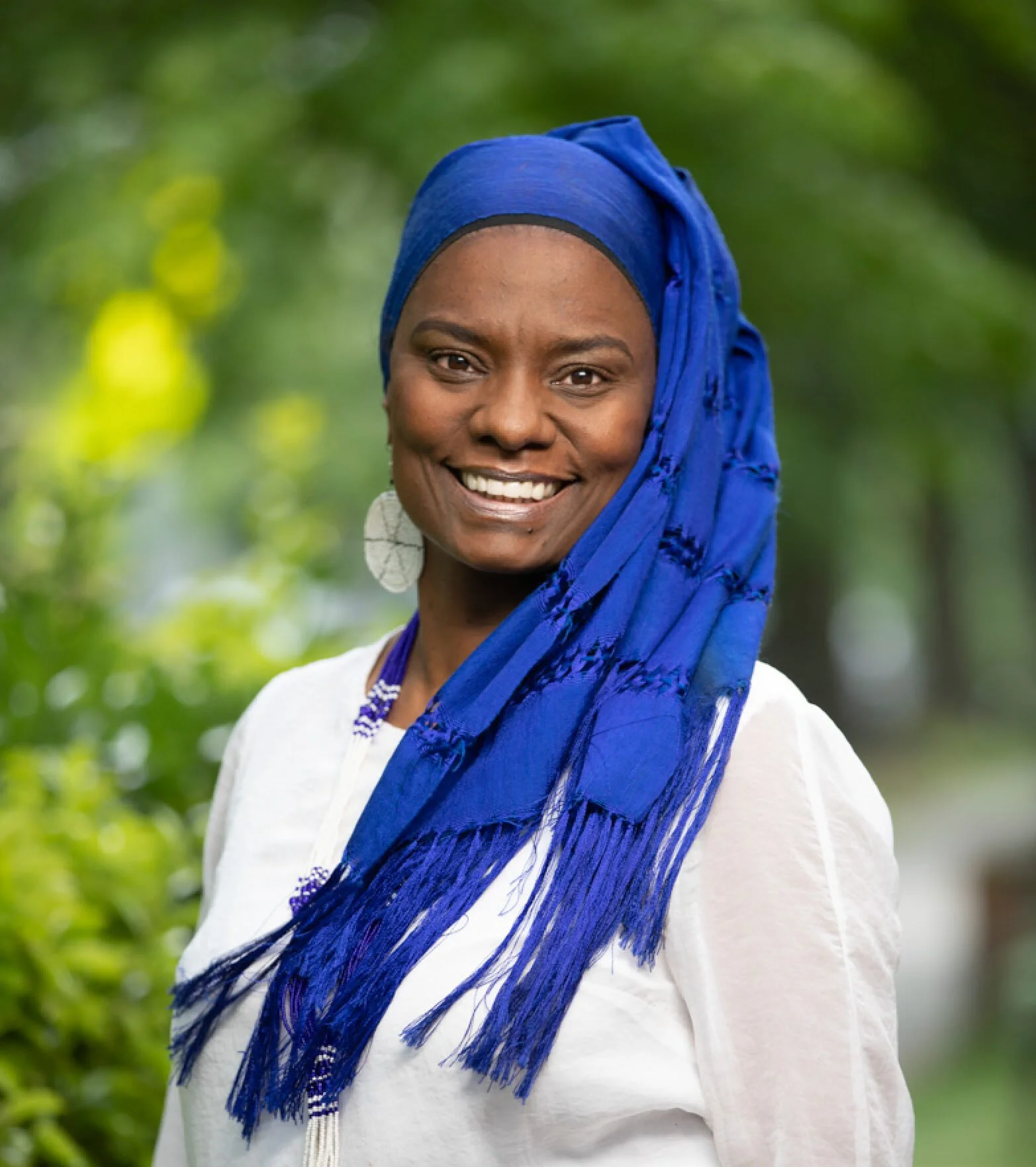 A smiling woman wearing a blue headwrap and white top outdoors with greenery in the background.