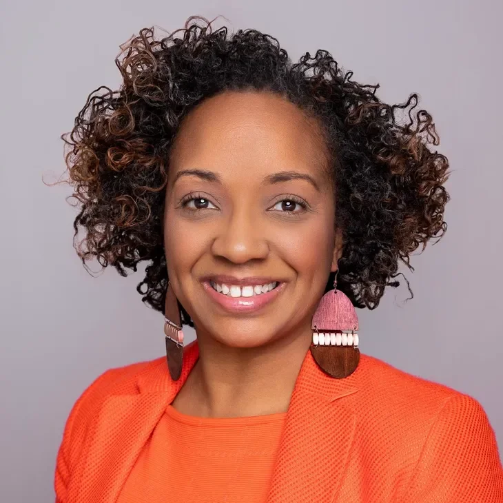 A woman with curly hair smiling, wearing a bright orange blazer and large pink and brown earrings.