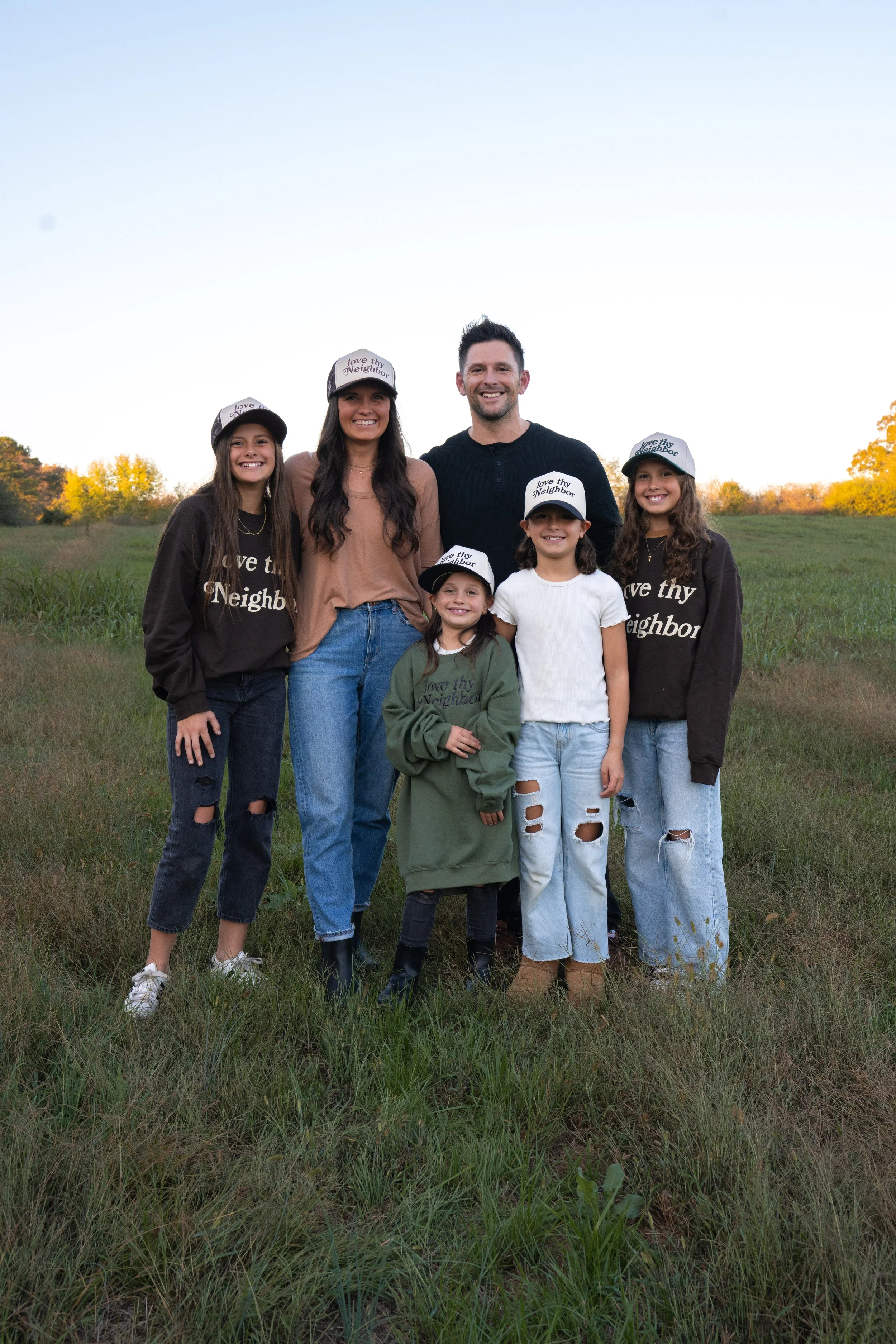 Family of six standing outside in a grassy field, smiling at the camera, wearing casual clothing and matching hats that say "Love Thy Neighbor."