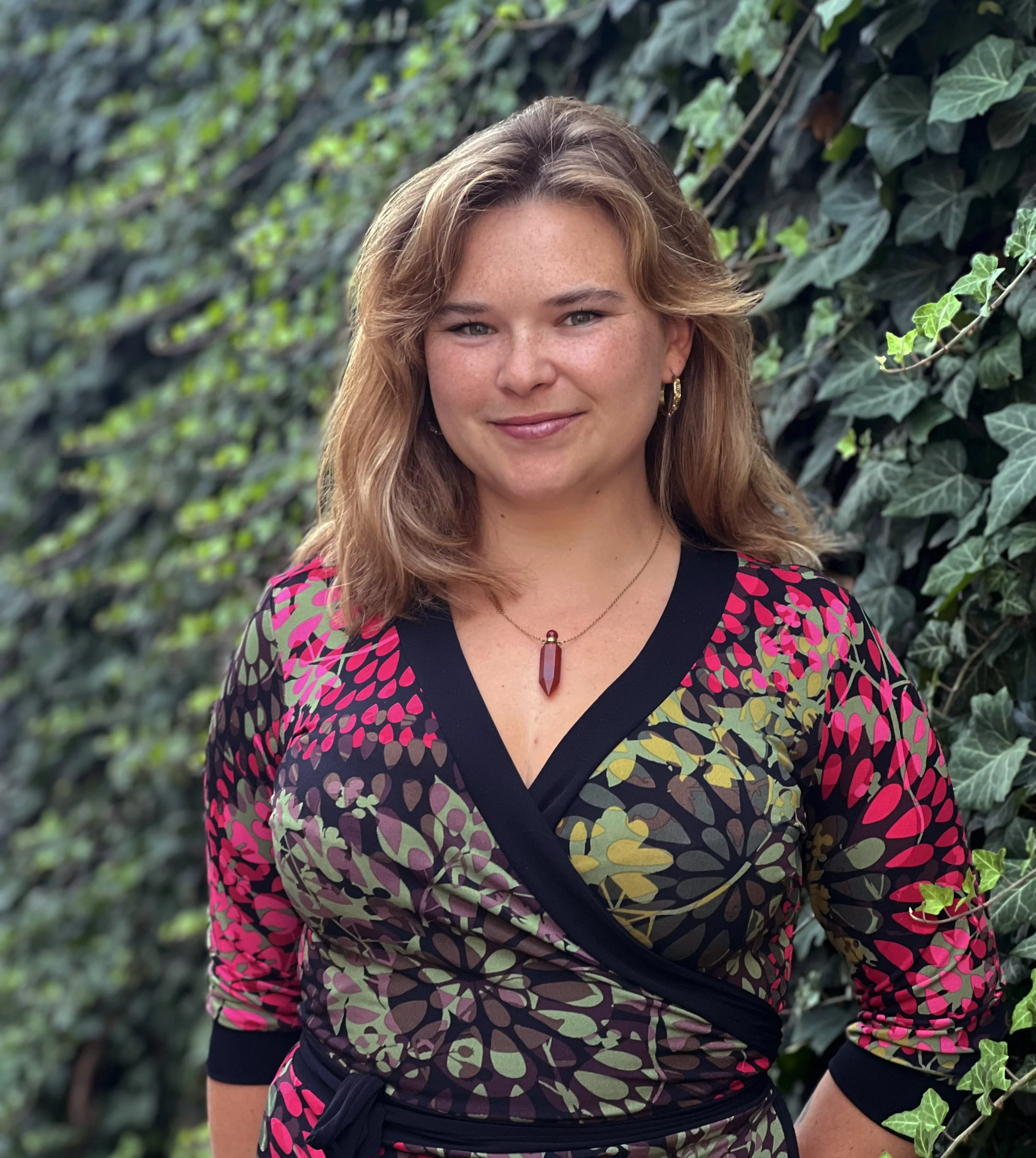 oman in a pink and black floral dress with curly blonde hair standing in front of ivy-covered wall, smiling.