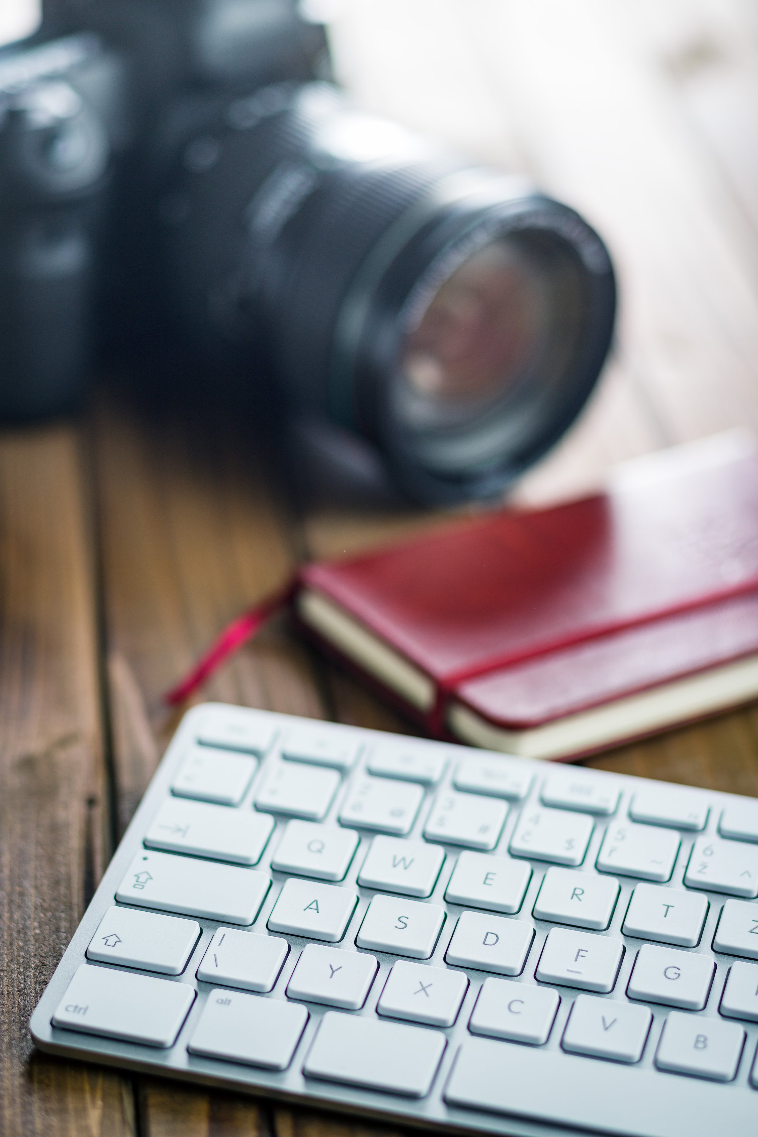 Close-up of a keyboard on a wooden desk with a blurred DSLR camera and a red notebook in the background.