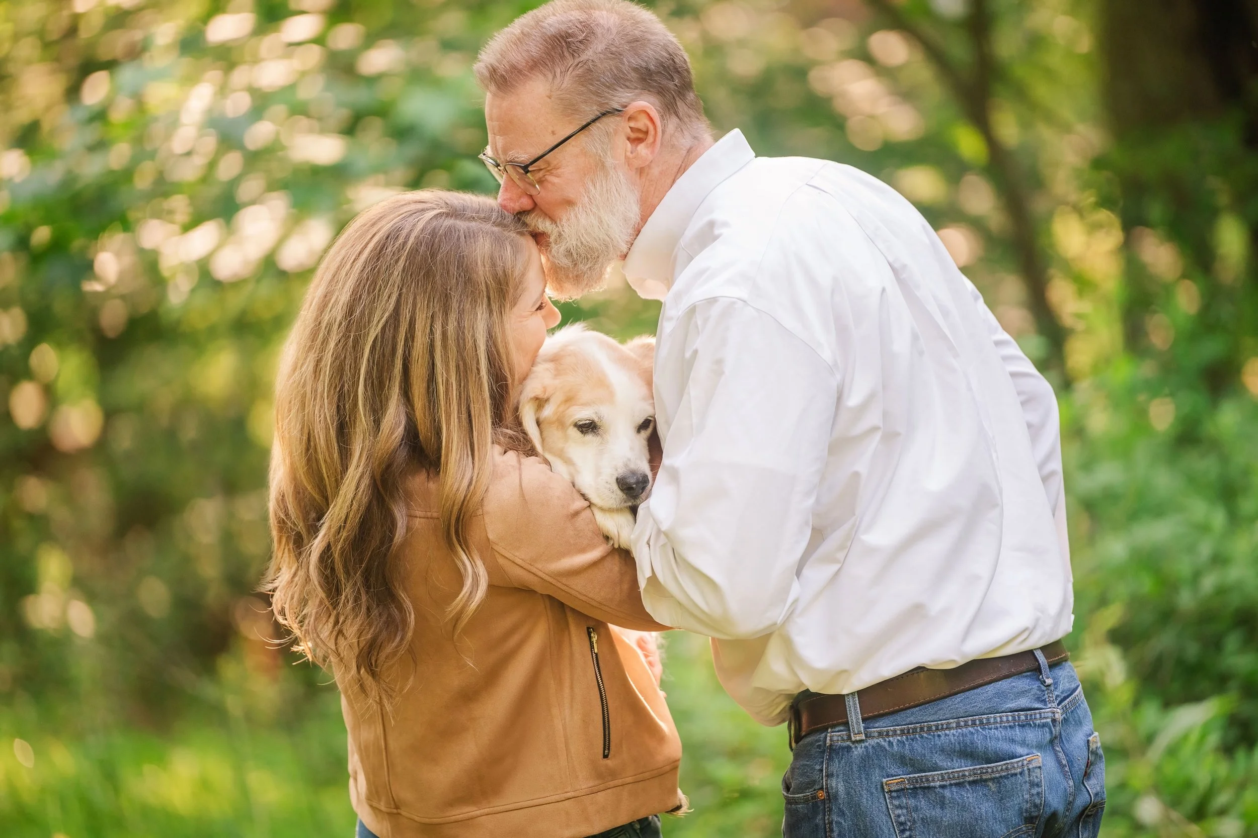 Man and woman with a small light-colored senior dog between them, soft outdoor light, trees in background. He's kissing her forehead and she's looking at the dog.