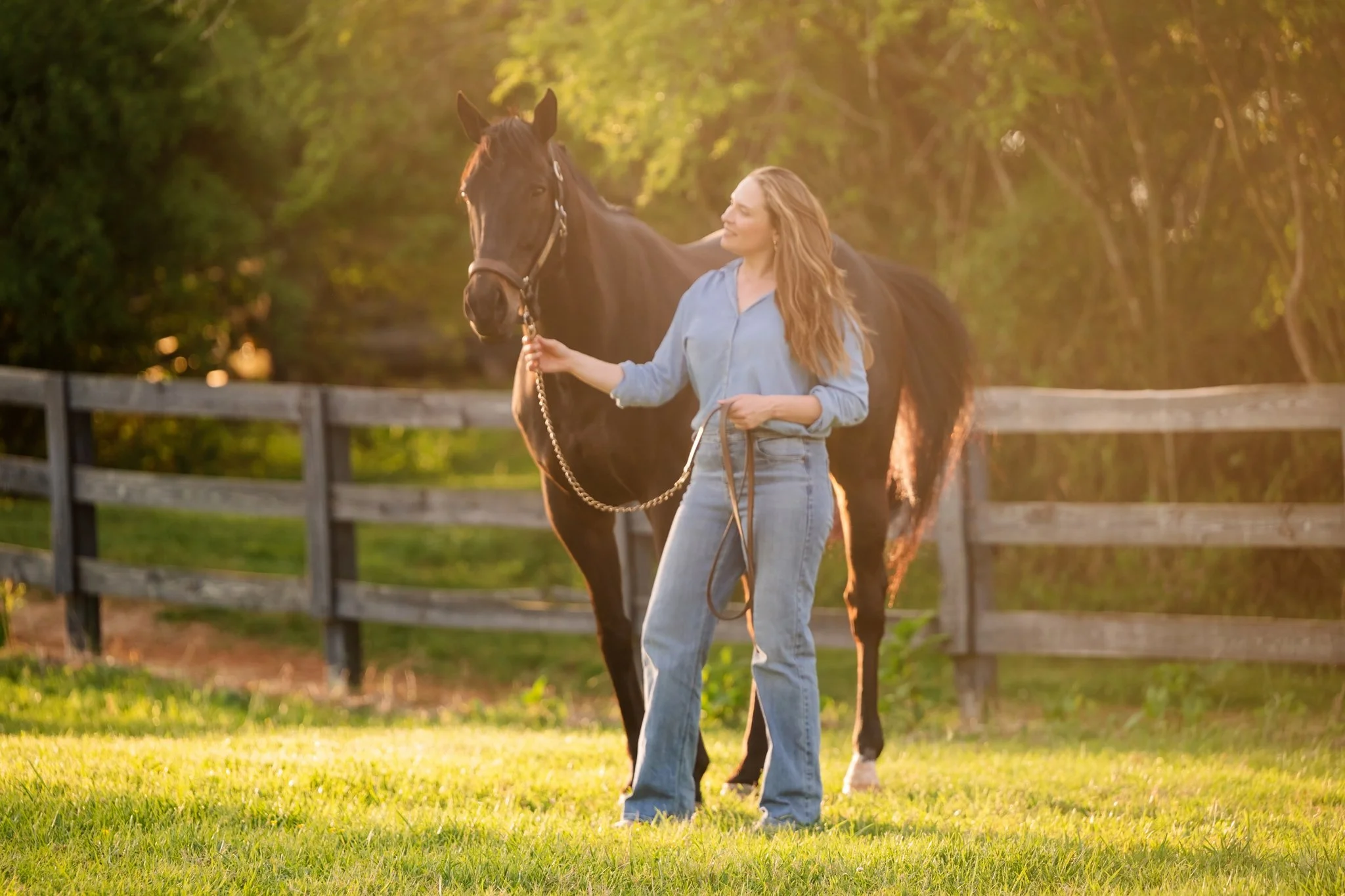 Woman in light blue denim holding lead rope and looking up lovingly at her black horse along a wooden fence line in stunning golden hour backlight at Arbor Hill Farm Canton Georgia