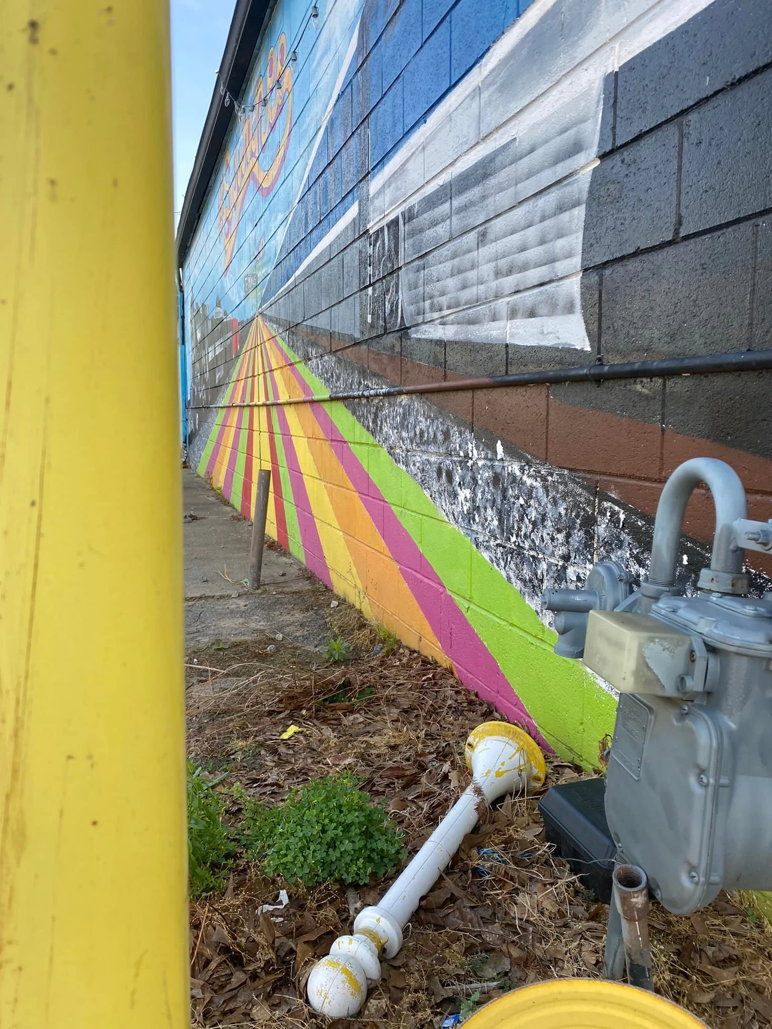 Ground-level phone photo looking down the side of the mural building. Yellow parking pole at left edge, colorful striped mural on the brick wall, gas meter and pipes at right, dead leaves and debris on the ground.