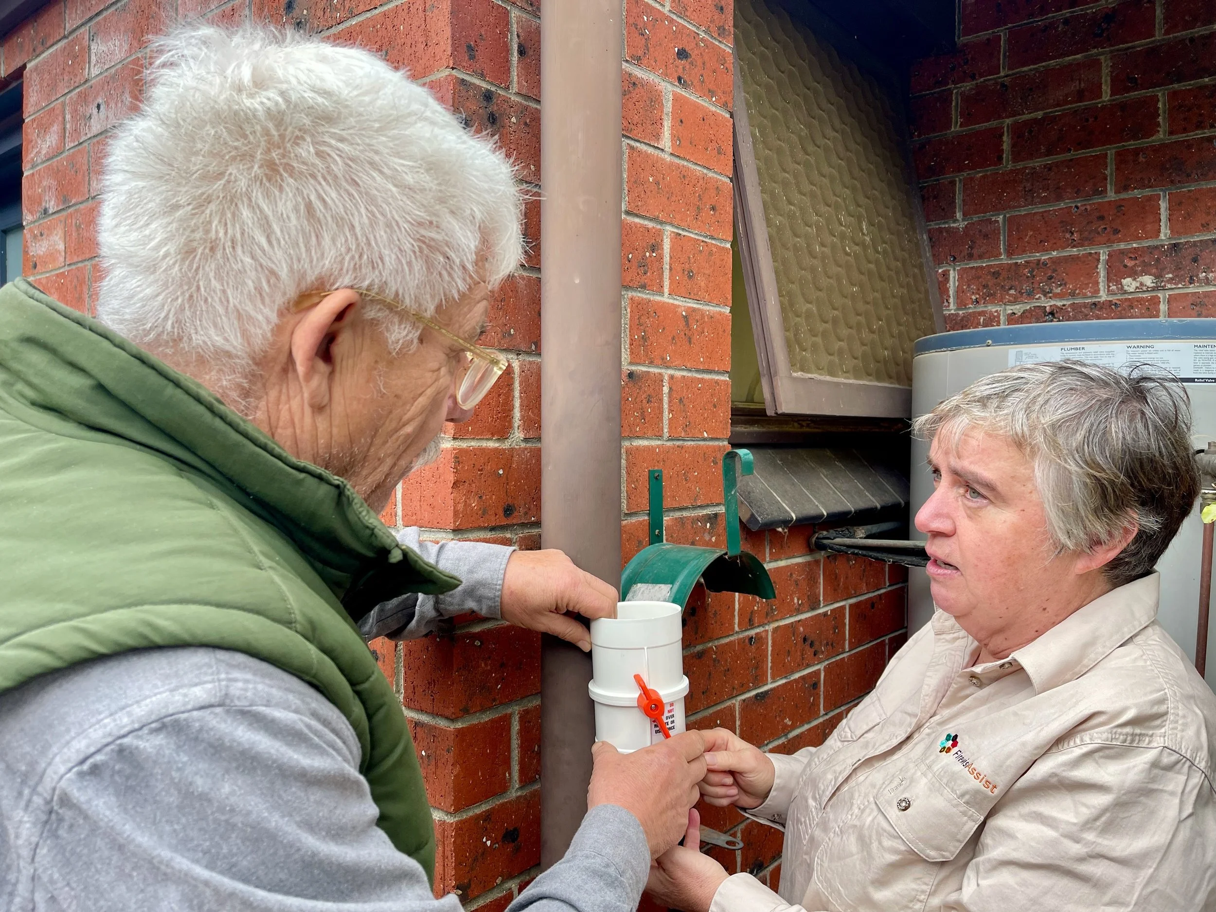 Two elderly men conversing outside a brick building; one man holds a white container with an orange twist-lock, the other man is reaching for it.